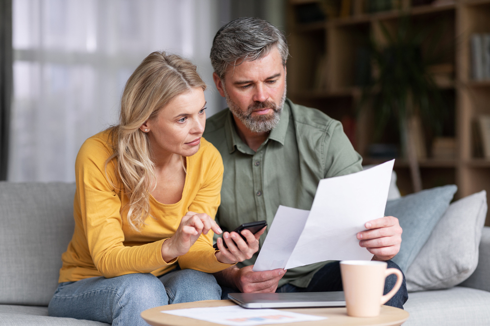 A man and a woman sit on a couch in Beach Park IL, reviewing a document together. The man holds the paper while the woman checks her smartphone. A laptop and a mug sit on the table as they discuss homeowners insurance options.
