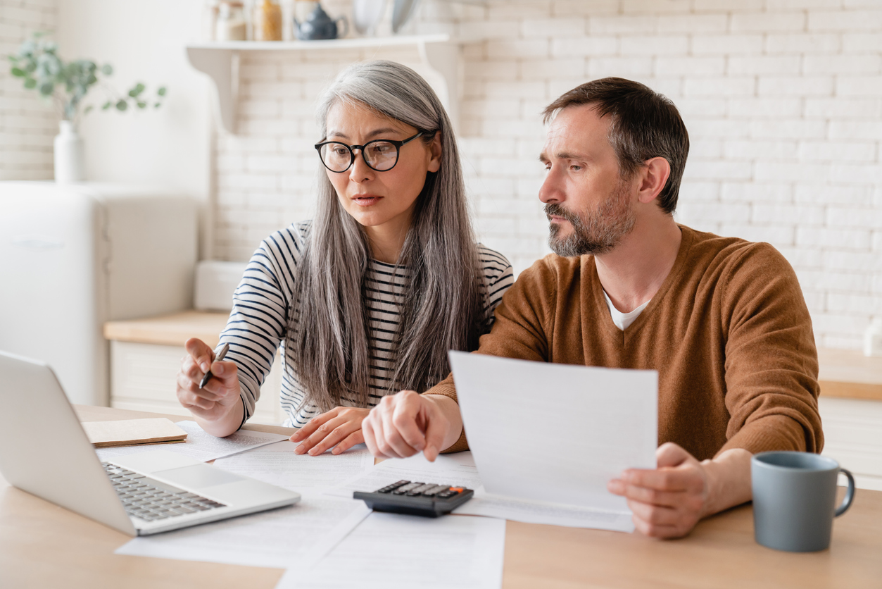 A woman and a man sit at a table with papers, a calculator, and a laptop in front of them, reviewing homeowners insurance options in Bellwood IL. The woman points at the laptop screen while the man holds a document. Both appear focused and serious.