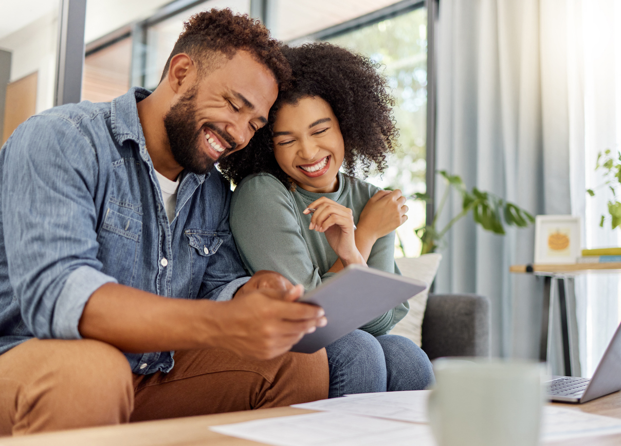 A man and woman sit closely together on a couch in their Belvidere, IL home, smiling at a tablet. Papers about homeowners insurance and a mug are on the table. They appear relaxed in a bright living room with large windows and indoor plants.