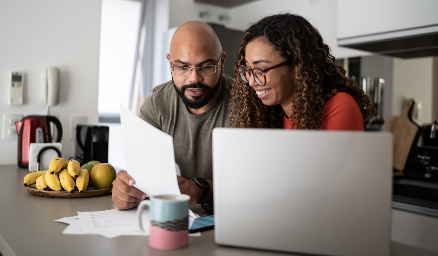 A man and woman sit at a kitchen counter in Belvidere IL, reviewing homeowners insurance documents together. A laptop, mug, and fruit are on the counter. Both appear focused, with the woman smiling at the papers.