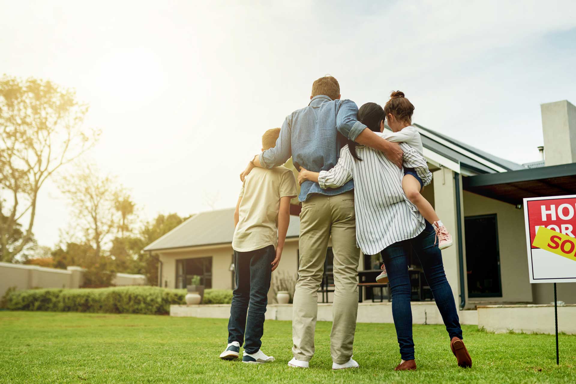 A family of four stands on a sunlit lawn in Bensenville IL, arms around each other, facing their modern house. A "For Sale" sign with a "Sold" sticker sits in the yard—a perfect moment to consider homeowners insurance.
