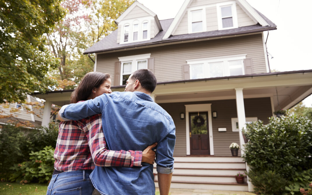 A couple stands arm in arm in front of their two-story Belleville IL home with beige siding and white trim, admiring it. The front yard has shrubs and trees—a perfect setting for new beginnings and reliable homeowners insurance.