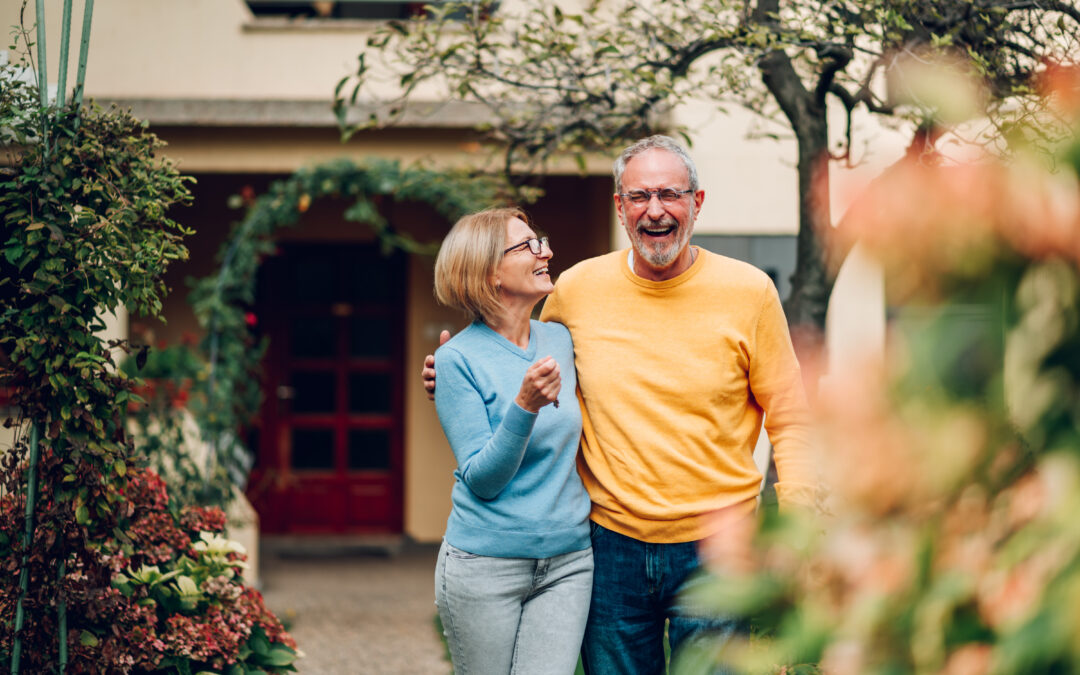 An older couple stands outside in a Bellwood, IL garden, smiling and holding each other. The woman wears a light blue sweater and glasses, while the man wears yellow. Greenery and flowers surround these happy homeowners enjoying their insurance-protected home.