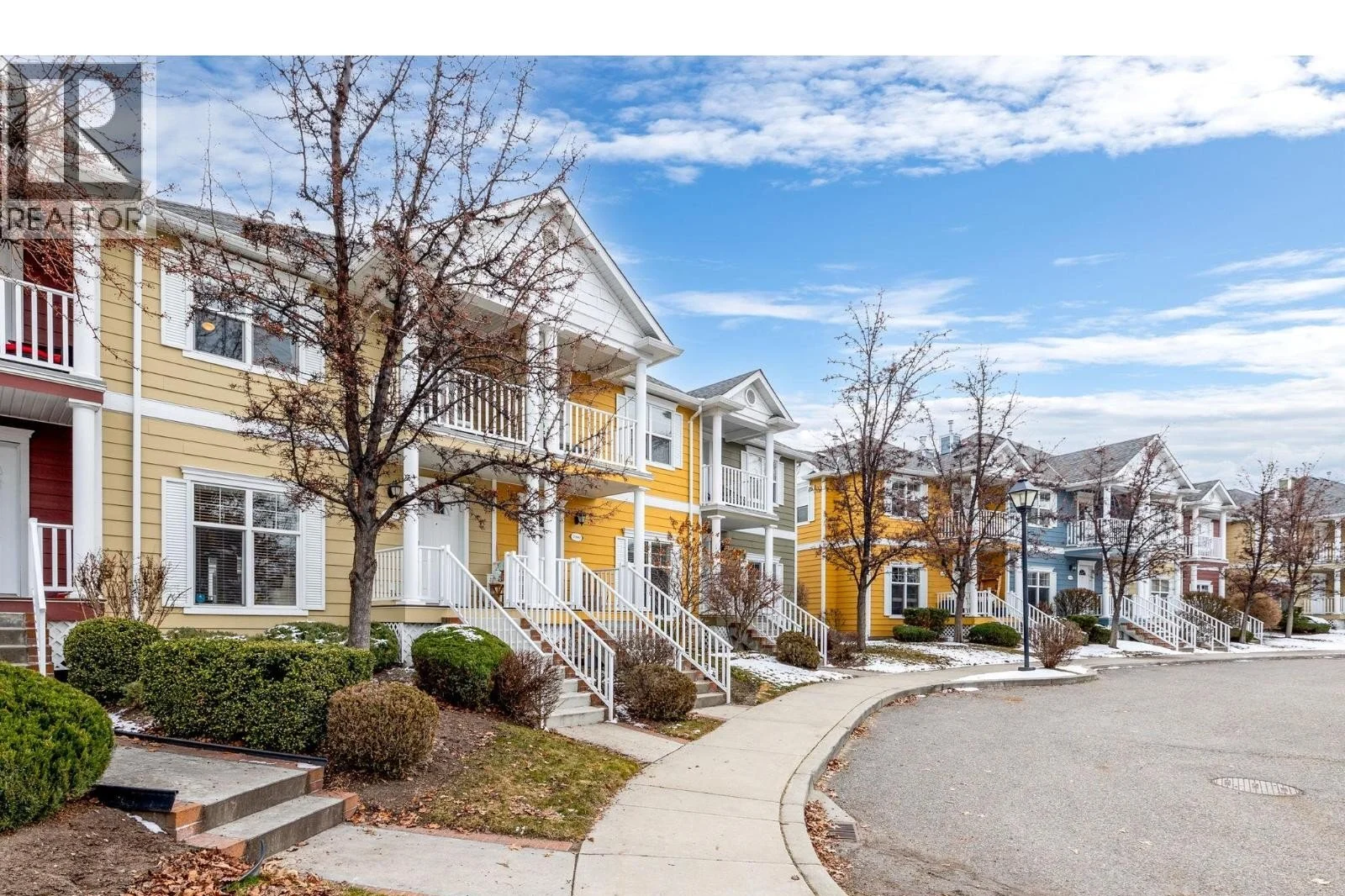 A row of two-story townhouse-style homes with yellow and blue siding, white railings, and small front porches lines a curved residential street in Bellwood IL, with bare trees, some snow on the ground, and homeowners insurance peace of mind.