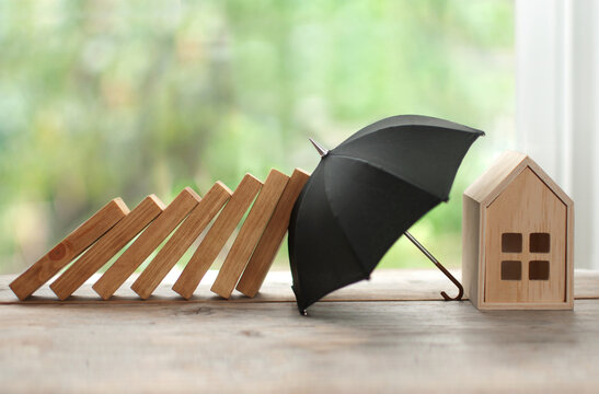 A row of wooden dominoes is falling toward a small house model in Belleville, IL, but a black umbrella—symbolizing homeowners insurance—stands between them, blocking the dominoes from hitting the house.