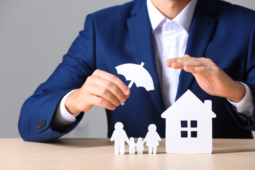 A person in a blue suit holds a paper umbrella over paper cutouts of a family and a house, symbolizing homeowners insurance protection in Bellwood IL, set against a neutral gray background.