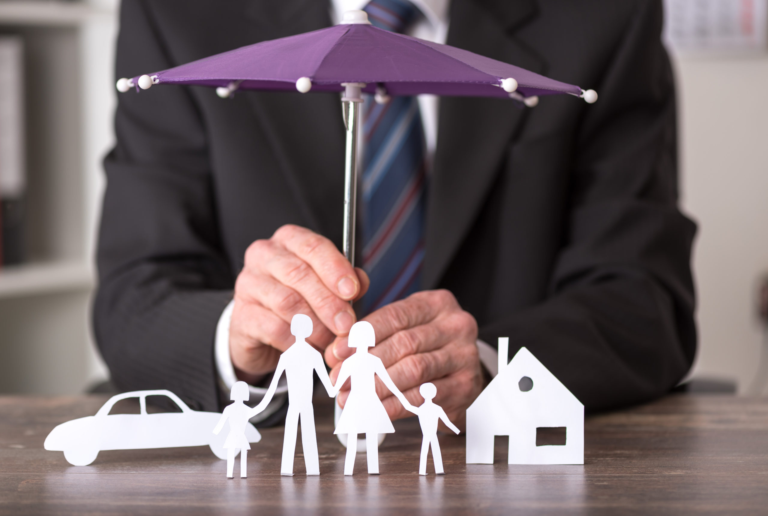 A person in a suit holds a small purple umbrella over paper cutouts of a family, a car, and a house, symbolizing homeowners insurance coverage in Belvidere IL.