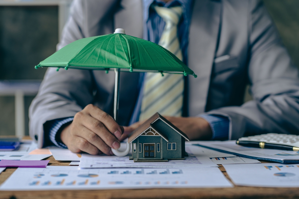 A person in a suit holds a green umbrella over a small model house on a desk covered with papers, charts, and a calculator, symbolizing homeowners insurance or property investment protection in Bensenville IL.