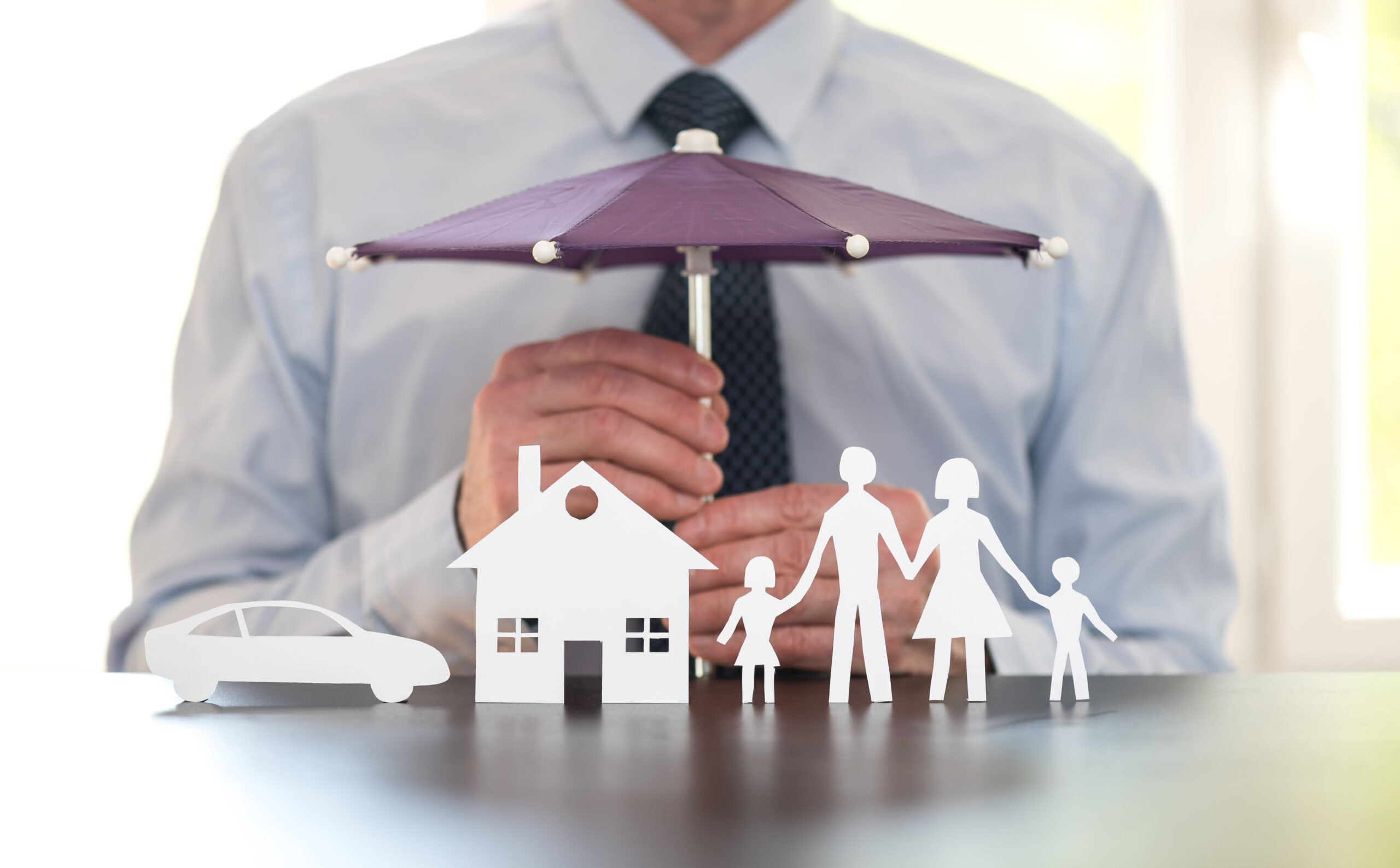 A man in a dress shirt and tie holds a purple umbrella over paper cutouts of a car, house, and family of four, symbolizing protection or insurance coverage such as Homeowners Insurance Blue Island.