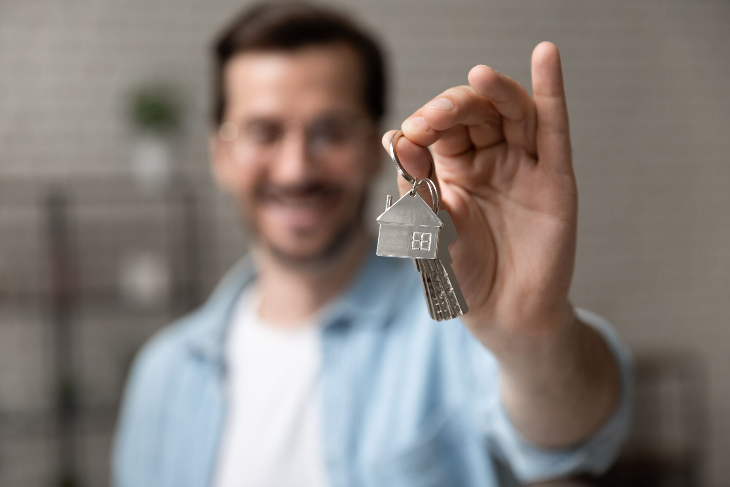 A person holds a set of house keys with a house-shaped keychain, extending them toward the camera. The person is smiling and wearing glasses, ready to settle in—with homeowners insurance Berwyn providing peace of mind in the blurred interior background.