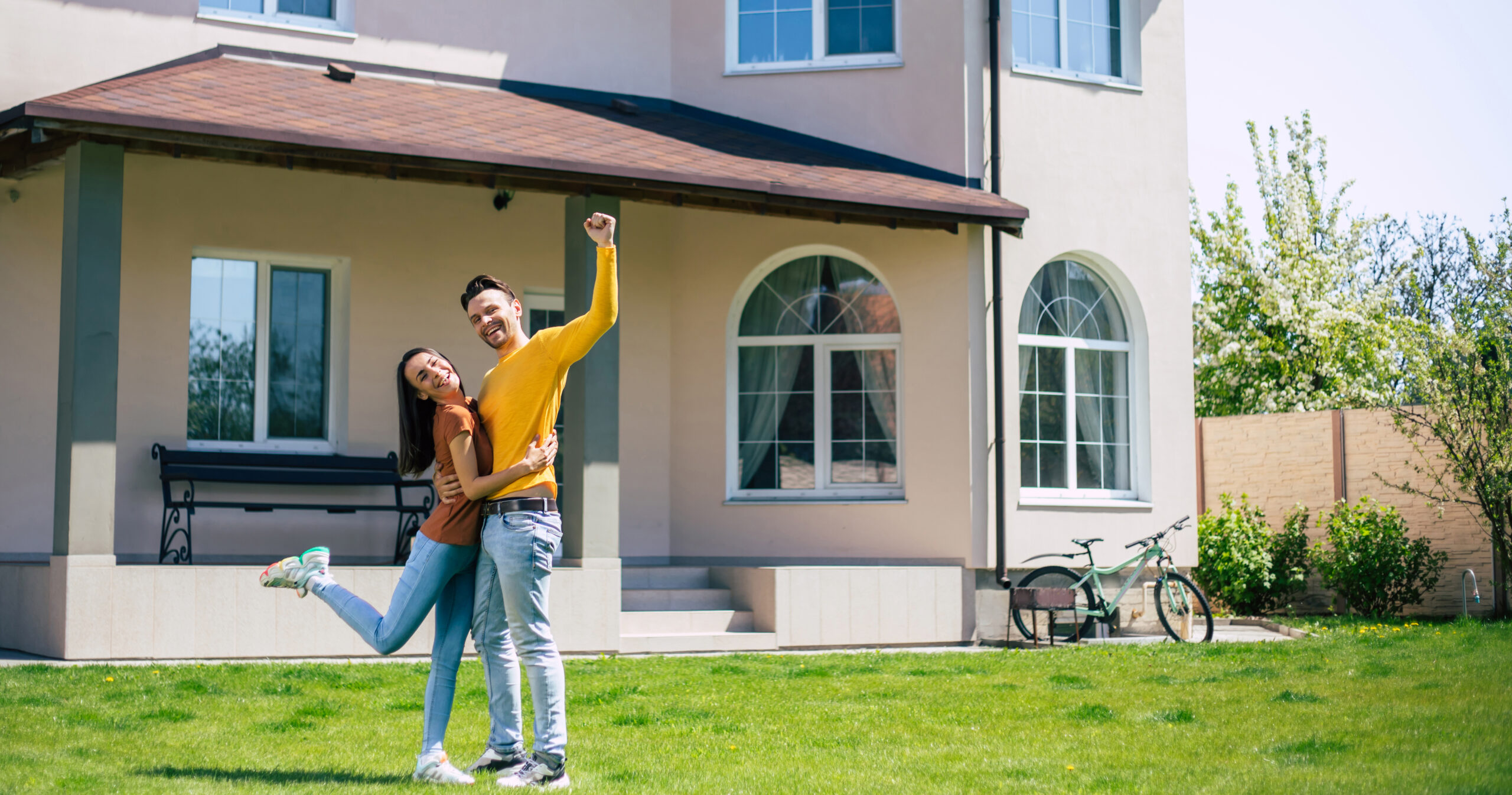 A smiling couple stands together on the lawn in front of a light-colored house. The man raises his arm in celebration while the woman lifts one leg—proud Bloomingdale homeowners insurance policyholders. A bicycle rests near a wooden fence.