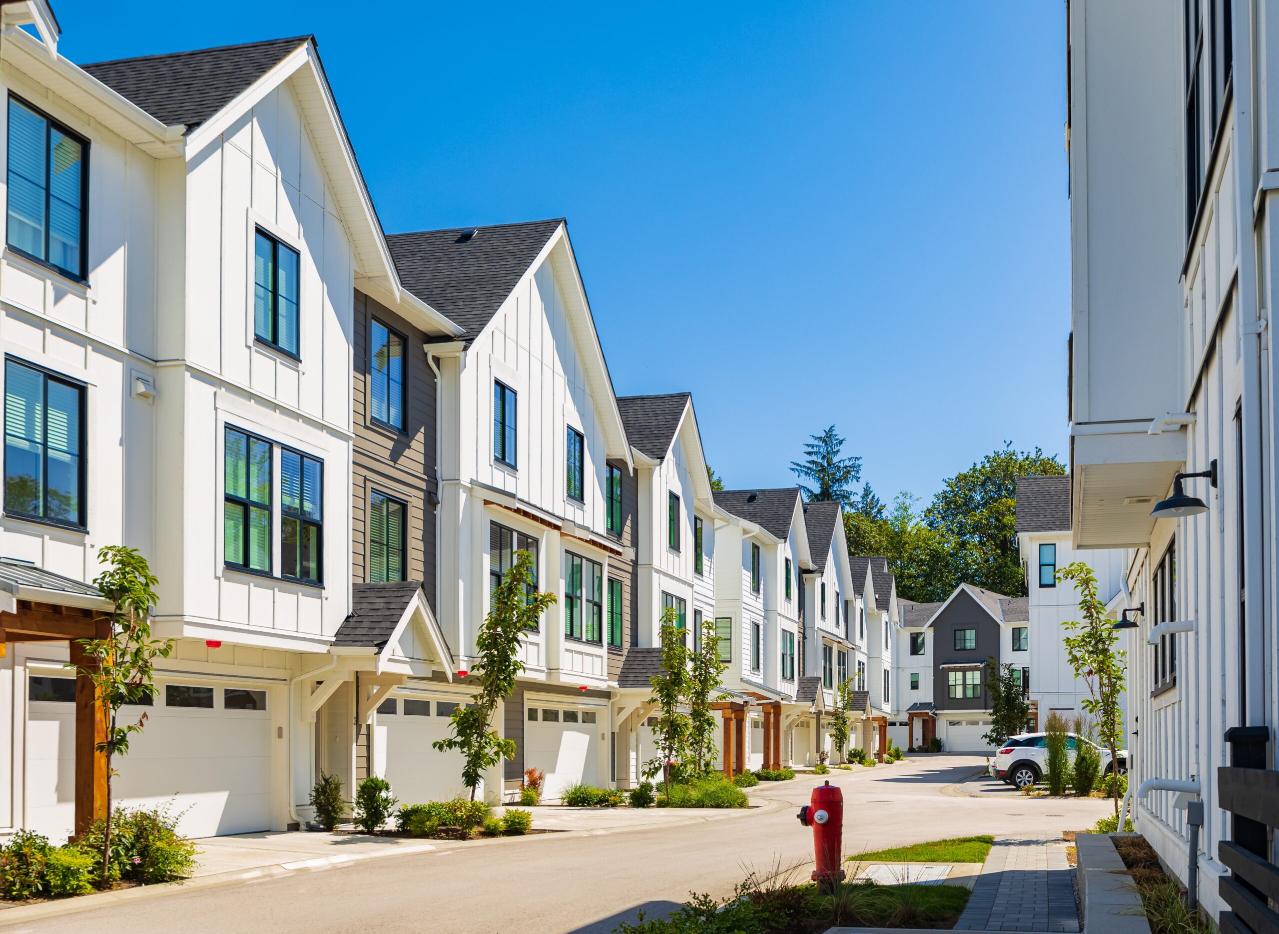 A row of modern, white multi-story townhouses with garages lines a clean street under a clear blue sky. Small trees dot the sidewalk as a red fire hydrant stands in the foreground—an ideal scene for Bloomingdale homeowners insurance coverage.