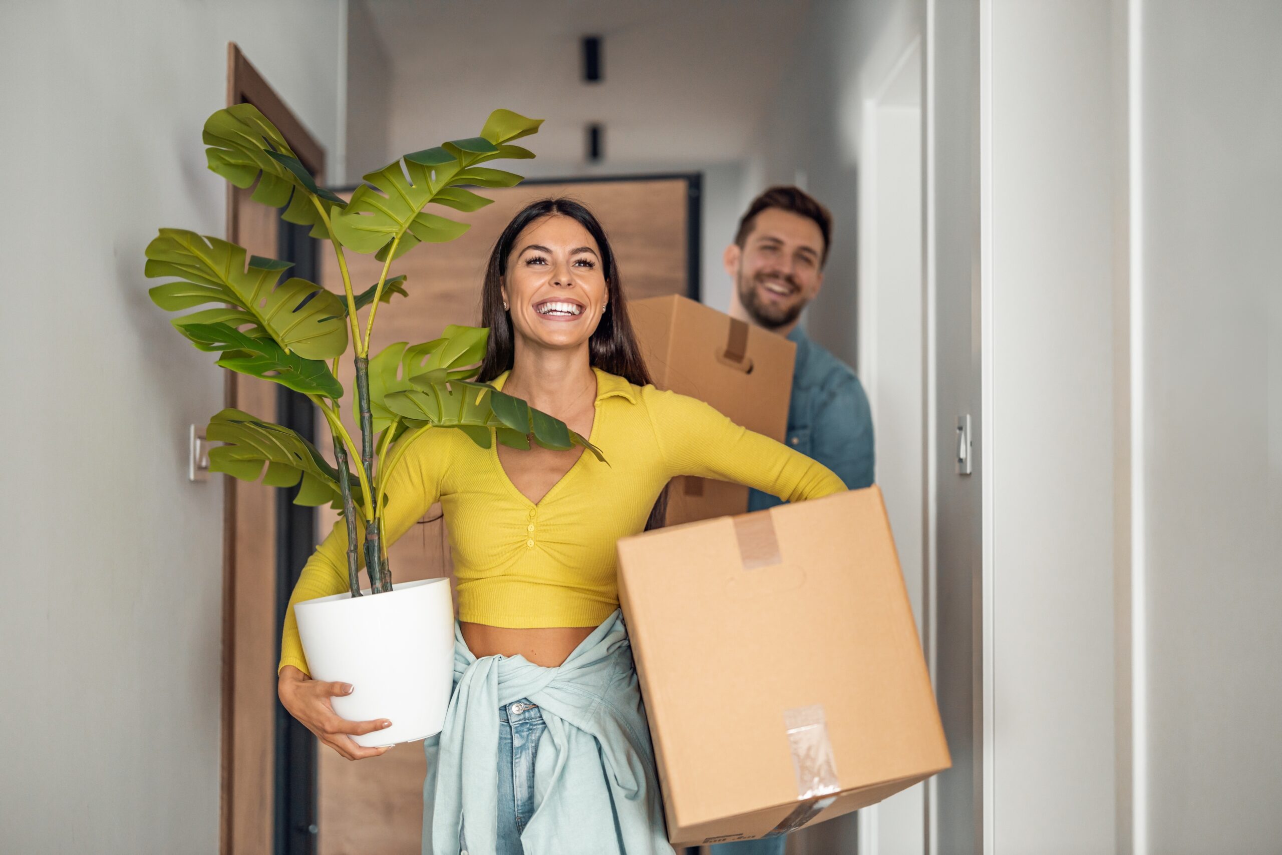 A woman carrying a potted plant and a cardboard box walks down a hallway, smiling, with a man behind her also holding a cardboard box—new beginnings made easier with Homeowners Insurance Blue Island.