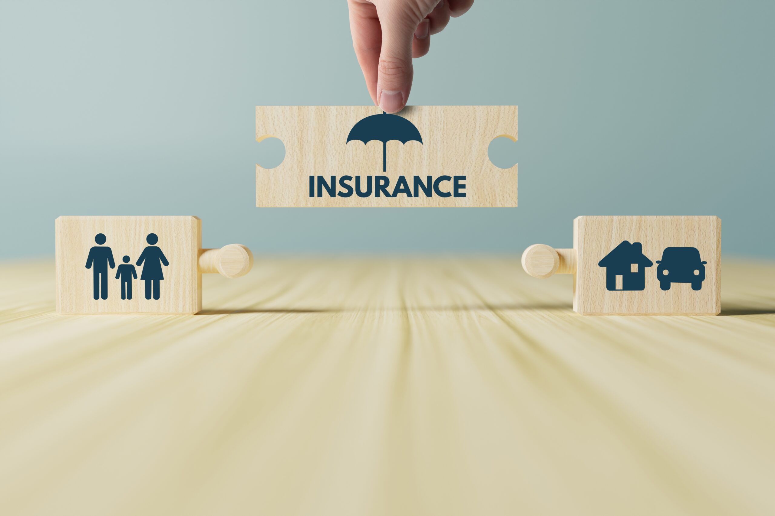 A hand places a puzzle piece labeled "INSURANCE" with an umbrella icon between two wooden blocks showing a family, house, and car—symbolizing protection for people and property through homeowners insurance in Bloomington, IL.