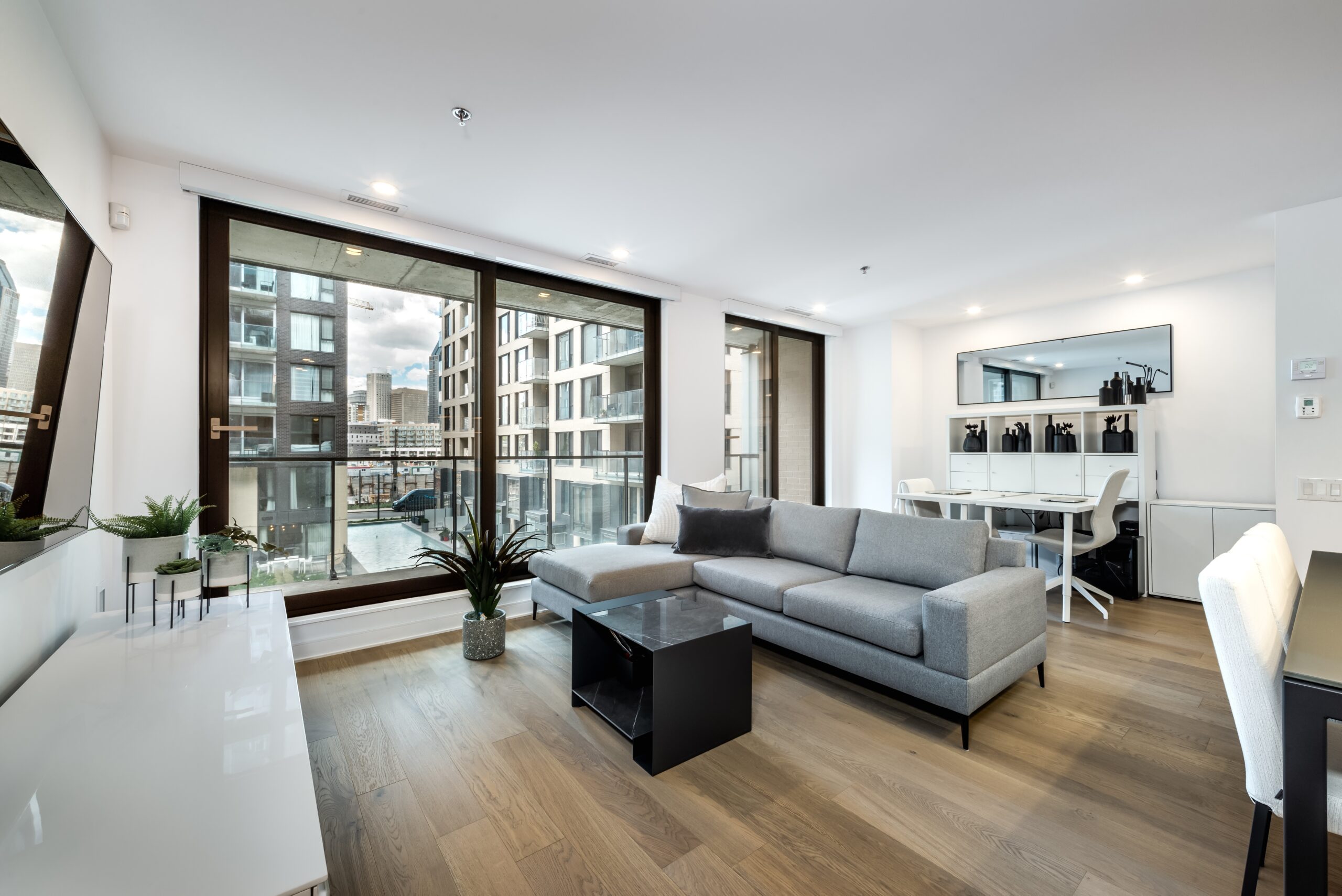A modern apartment living room with large windows, a gray sectional sofa, a black coffee table, a dining area with a white table and chairs, wood flooring, and views of nearby buildings—ideal for those seeking Homeowners Insurance Bolingbrook IL.