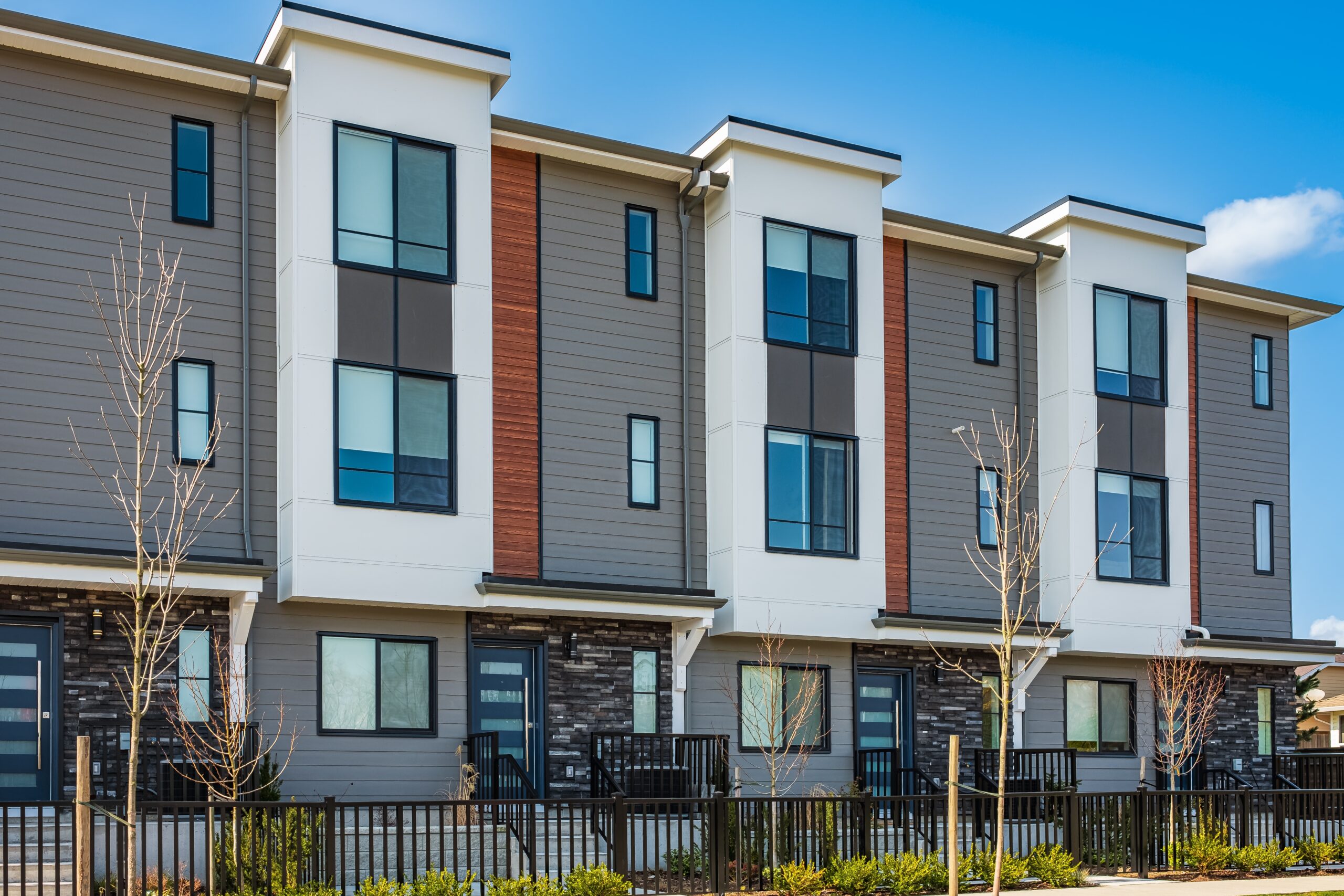 Modern three-story townhouse building with gray and white exterior, large windows, and small fenced front yards. Newly planted trees and shrubs are visible—ideal for residents seeking Bloomingdale homeowners insurance under a clear blue sky.