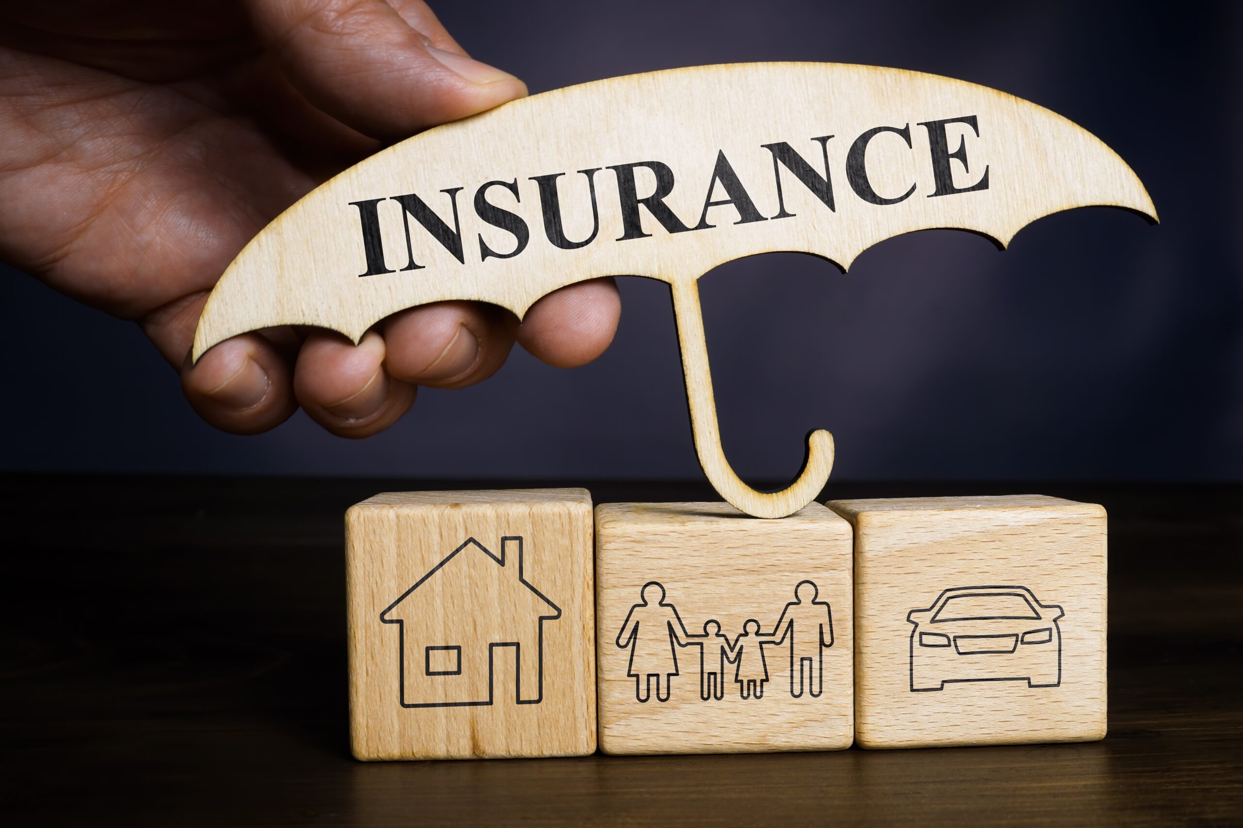 A hand holds a wooden umbrella labeled "INSURANCE" above three wooden blocks showing icons of a house, family, and car, symbolizing comprehensive coverage like Bloomingdale homeowners insurance.