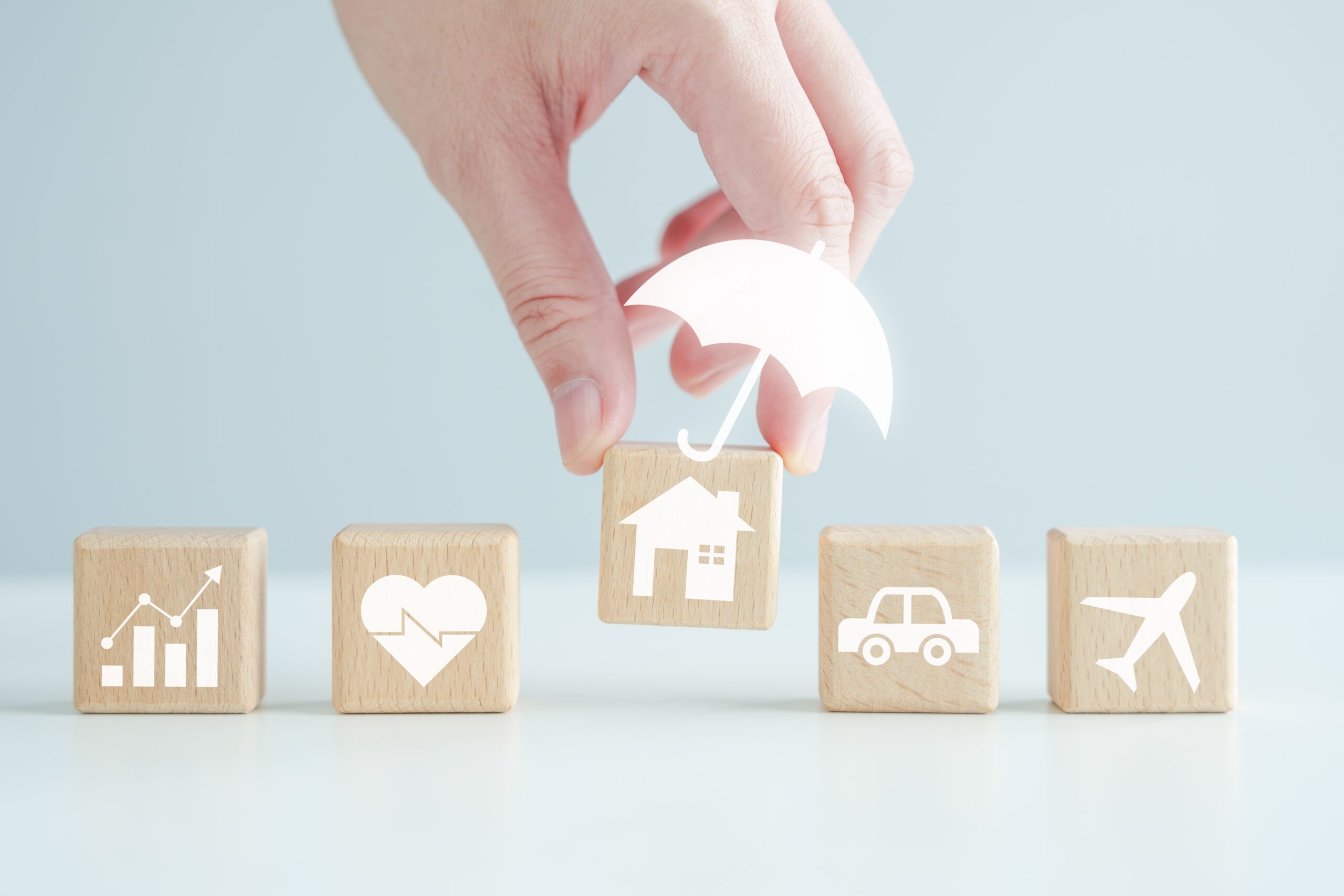 A hand places a wooden block with a house icon and a white umbrella above it among blocks for growth, health, car, and airplane—an image symbolizing the diverse coverage options in Homeowners Insurance Bolingbrook IL.