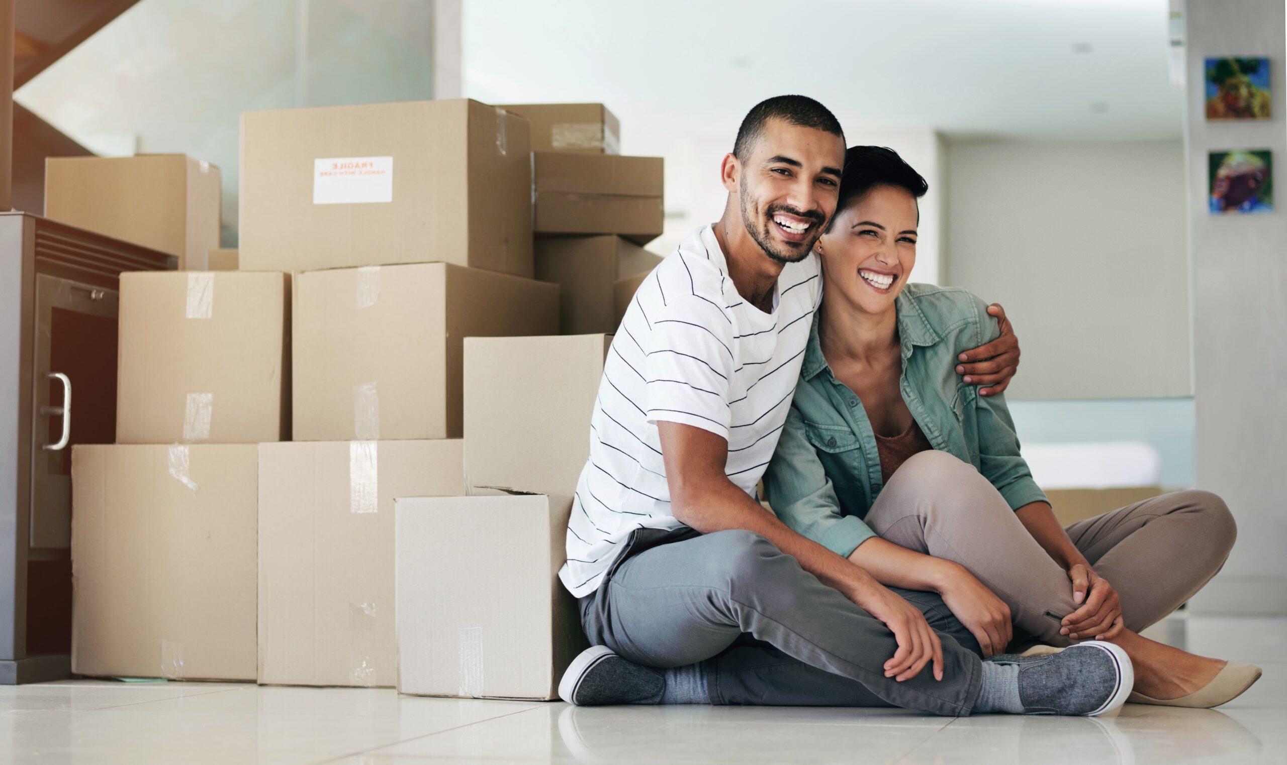 A smiling couple sits on the floor in front of a stack of cardboard moving boxes in a brightly lit room, celebrating their new space and peace of mind with Homeowners Insurance Blue Island. The man has his arm around the woman as they pose together.