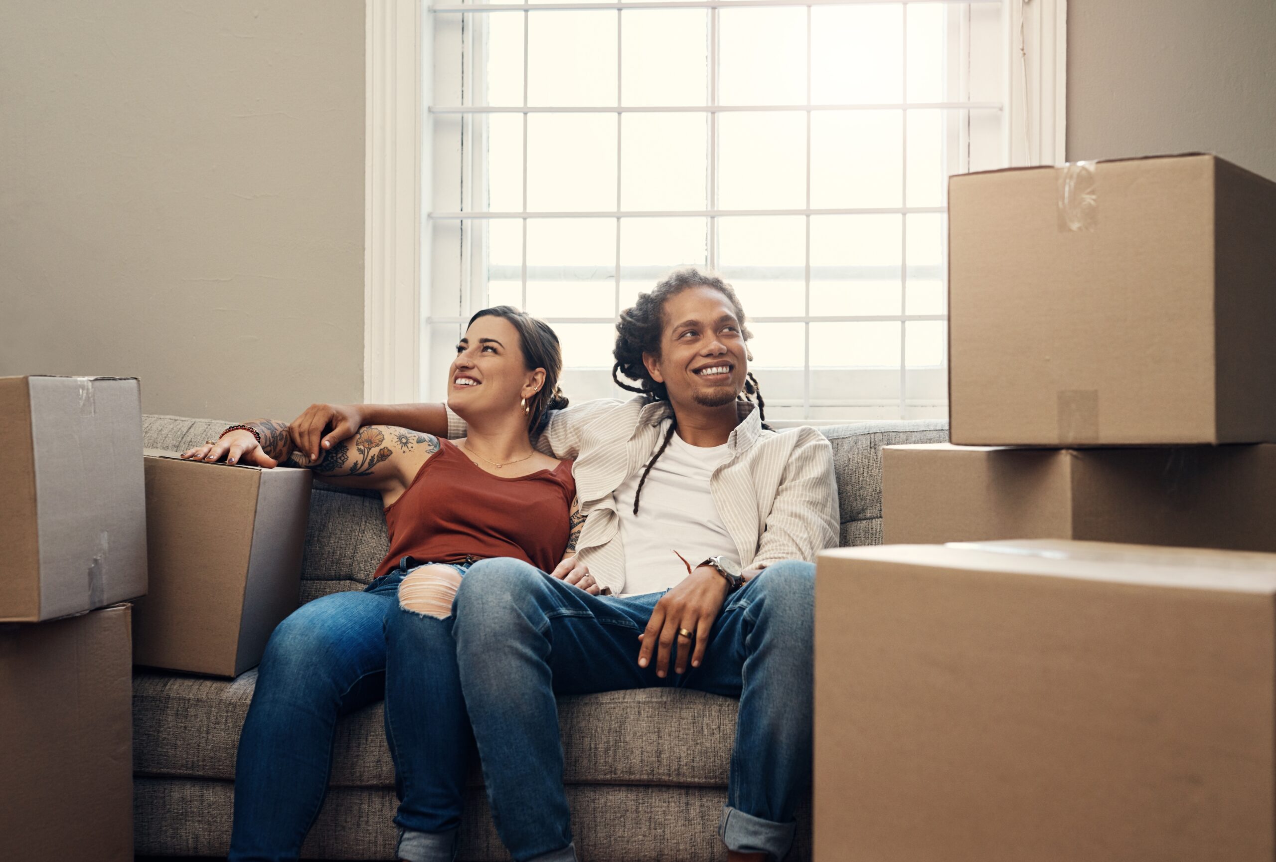A couple sits on a couch surrounded by cardboard moving boxes, smiling as they look off to the side. Sunlight fills the room through a large window, capturing the excitement of new beginnings protected by Addison homeowners insurance.