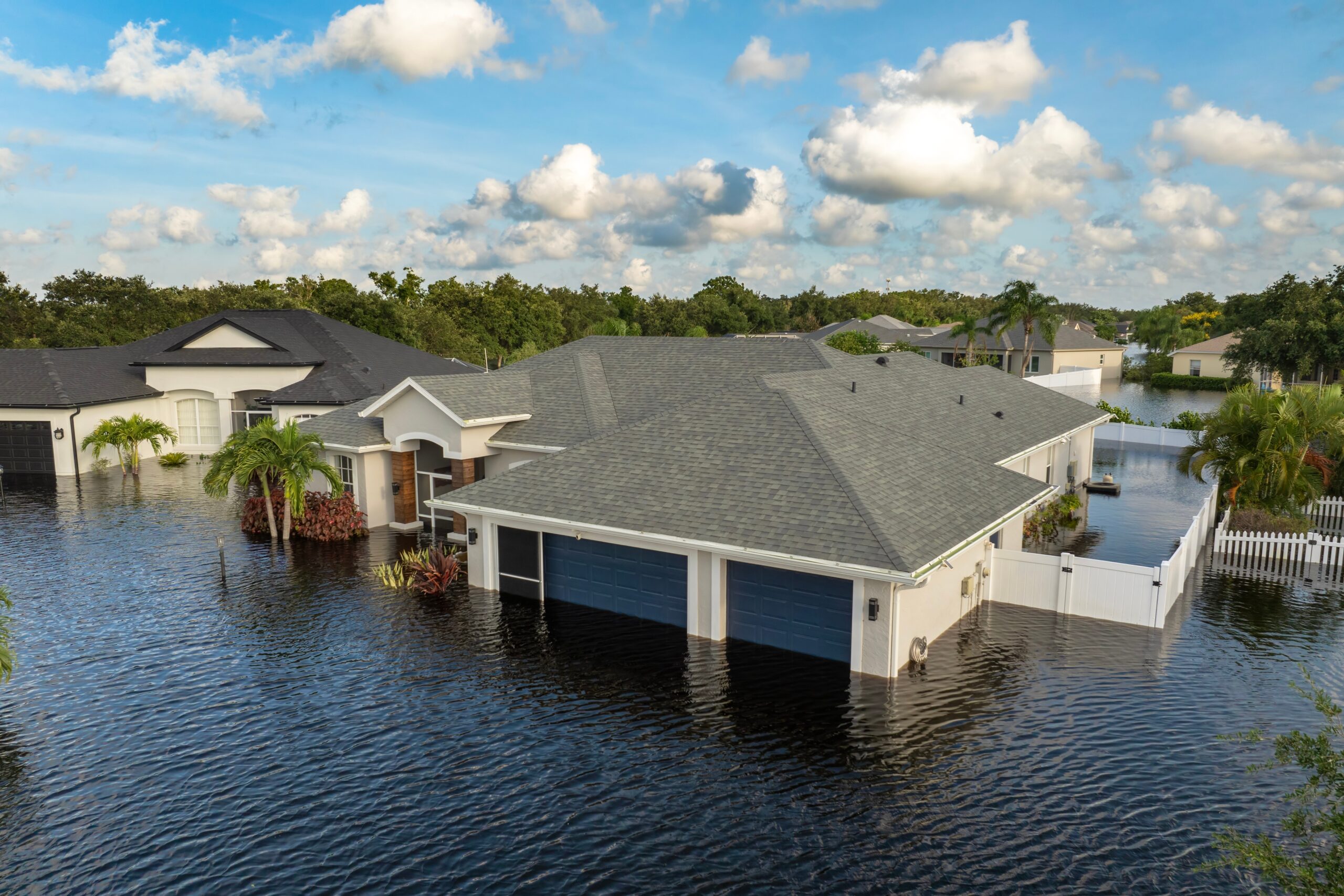 Single-story houses in Blue Island are surrounded by floodwater reaching halfway up garages and yards. Palm trees and other vegetation are partially submerged, highlighting the importance of homeowners insurance Blue Island residents rely on.