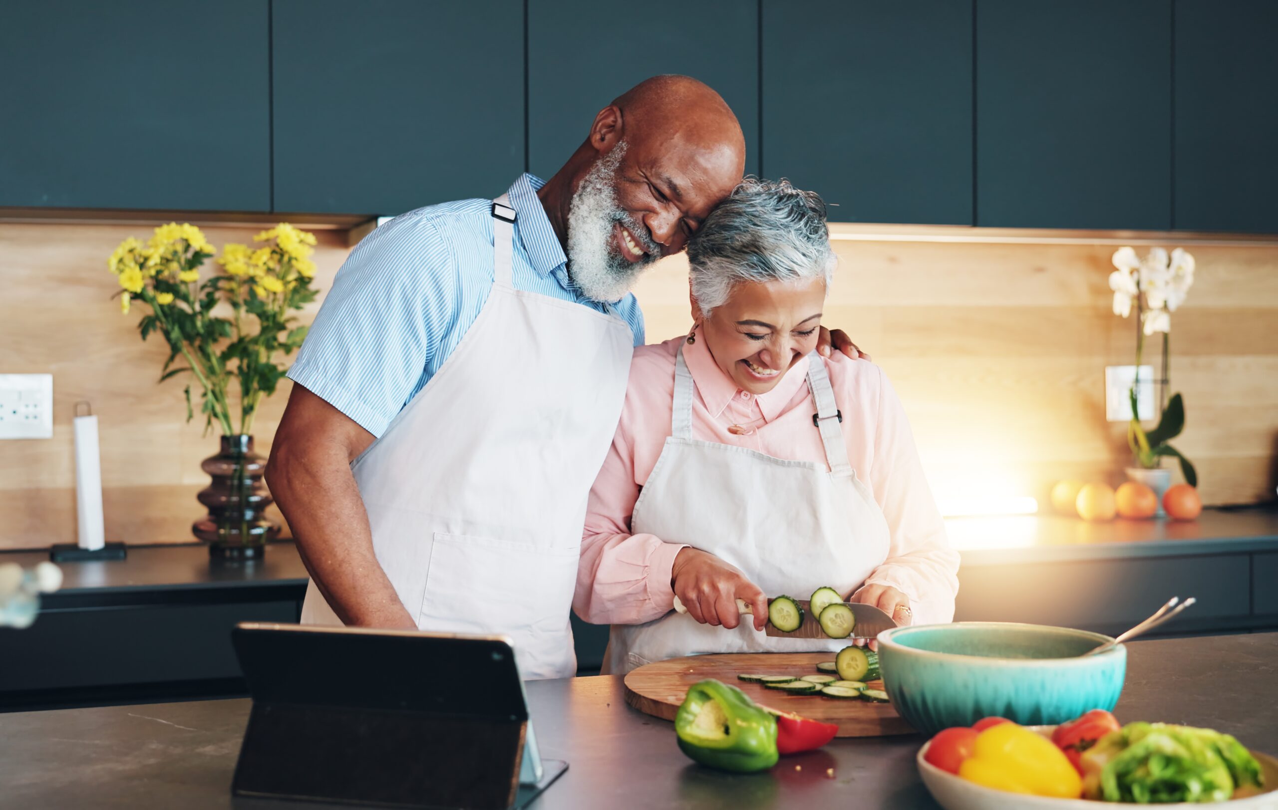 An older couple wearing aprons stands in a modern kitchen. As they prepare fresh vegetables together, a tablet sits on the counter—perhaps they're checking up on their Homeowners Insurance Bolingbrook for peace of mind in their cozy home.