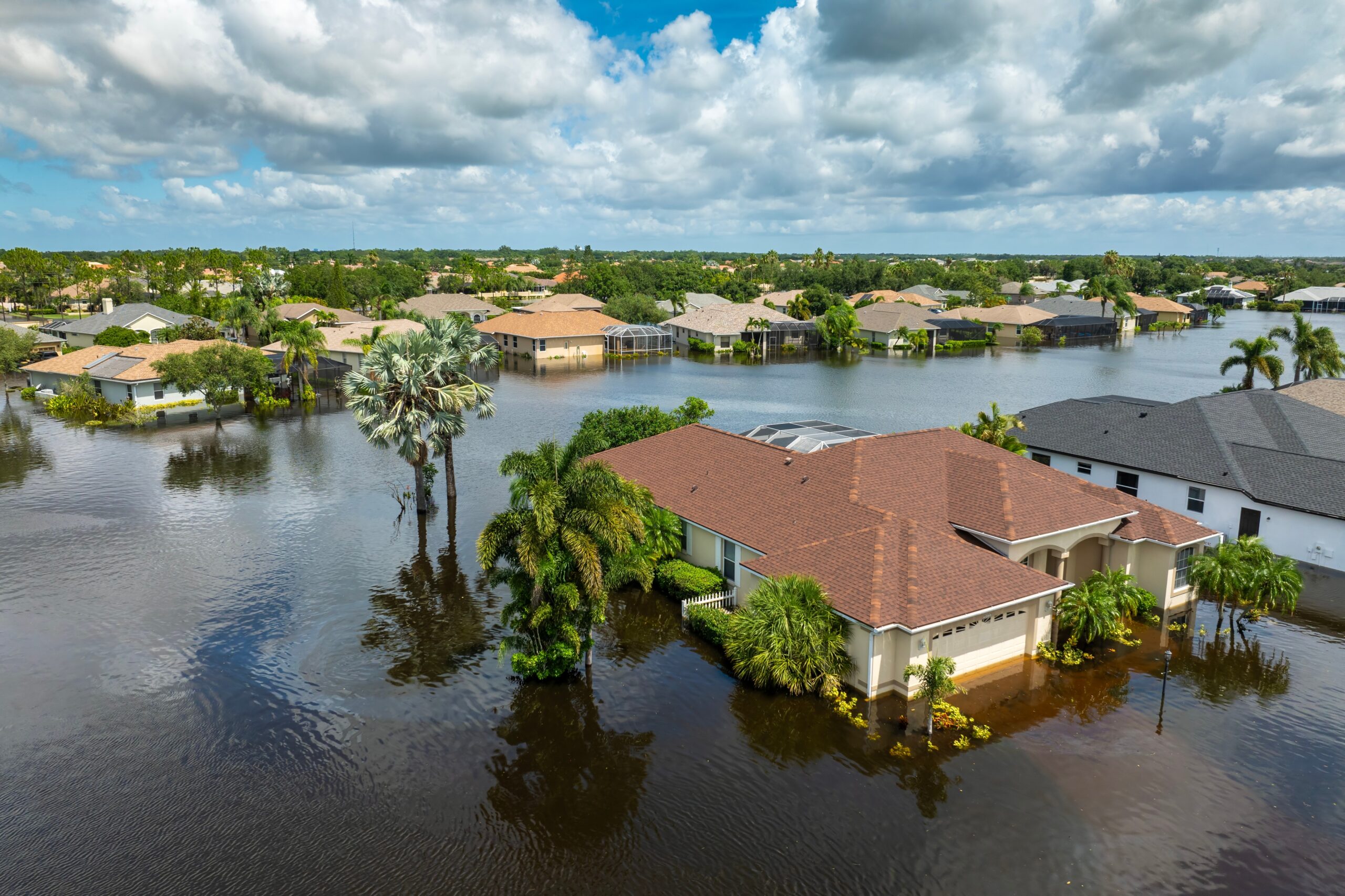 A suburban neighborhood is flooded, with water surrounding houses, trees, and lawns. The sky is partly cloudy, and the floodwater reflects the buildings and vegetation—highlighting the importance of Bloomingdale homeowners insurance.