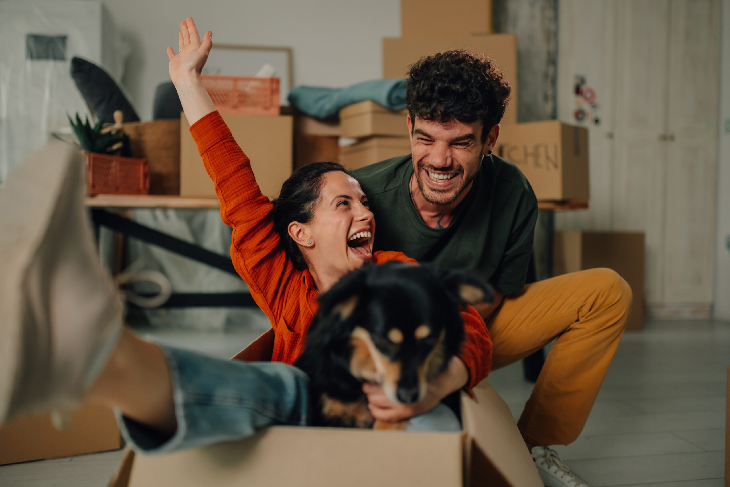 Two people smiling and laughing sit together in a cardboard box, holding a small black and brown dog. Moving boxes and household items are visible in the background—capturing the joy of new beginnings with Bloomingdale homeowners insurance.