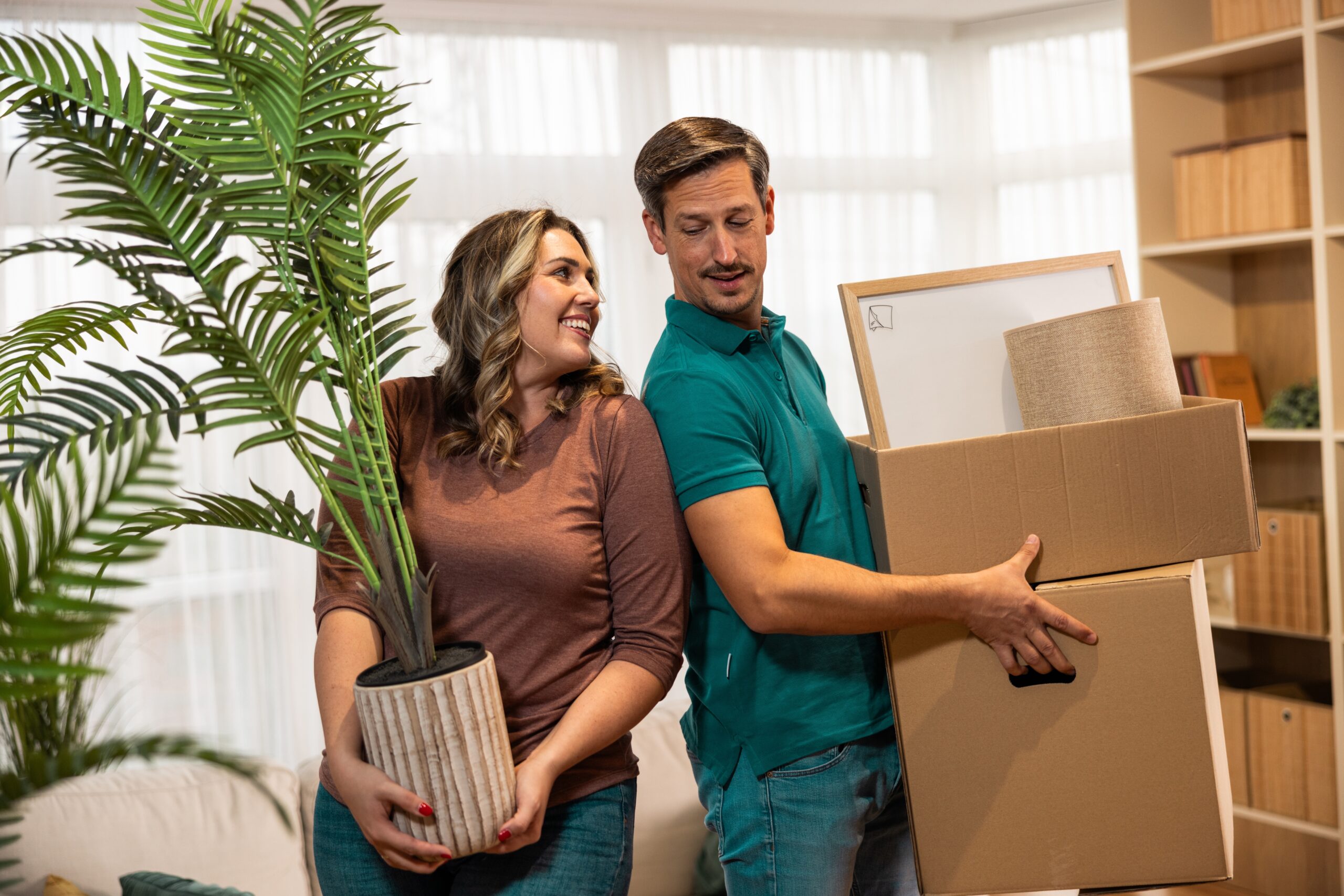 A woman holds a large potted plant while a man carries stacked cardboard boxes and a lampshade—new beginnings indoors. Discover peace of mind with homeowners insurance Berwyn as they settle in near sunlit windows and shelves.