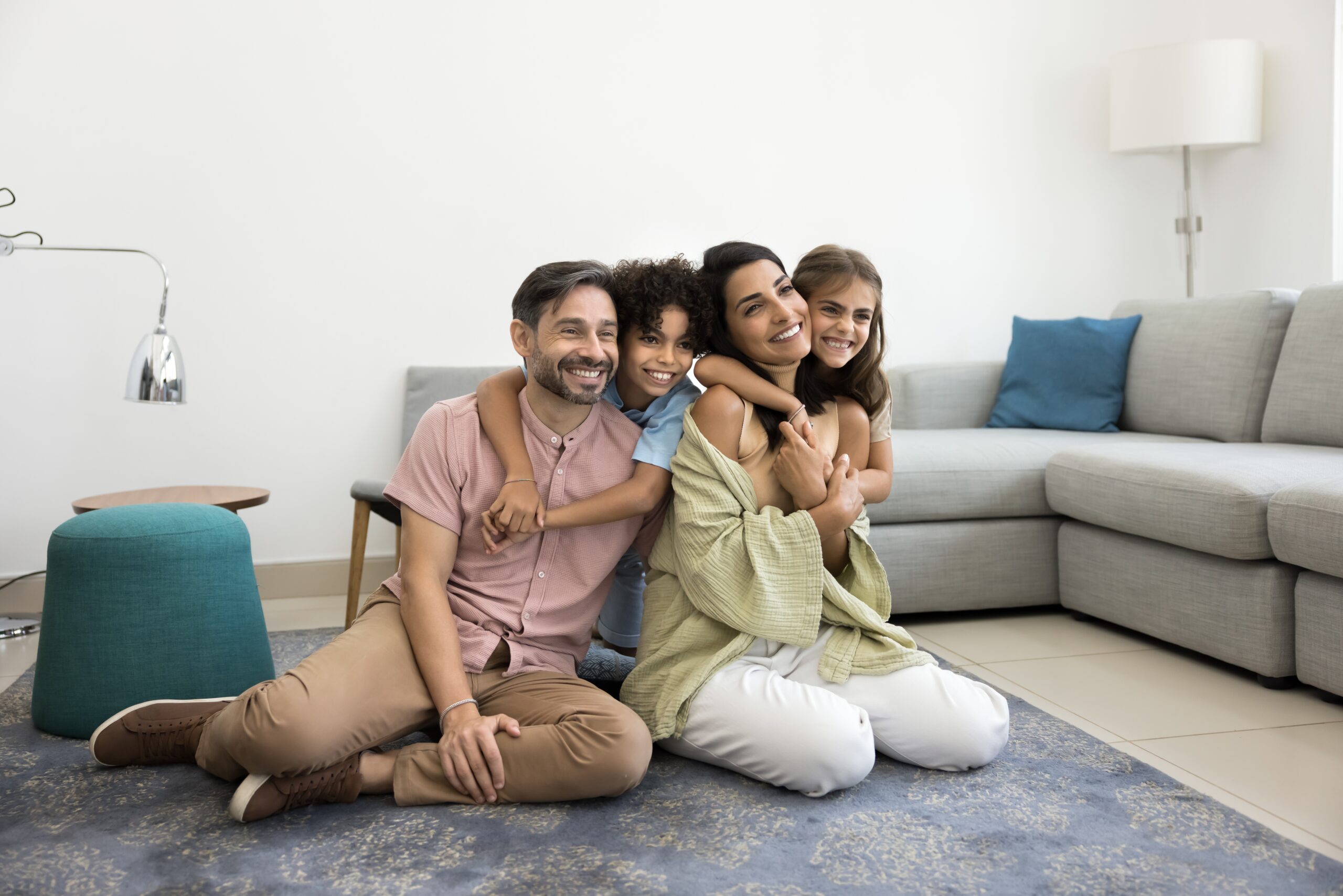 A man, woman, and two children sit on a blue rug in a living room, smiling and hugging each other. A gray sofa, blue cushions, and a green ottoman are visible—true comfort for those with homeowners insurance Bloomington IL.