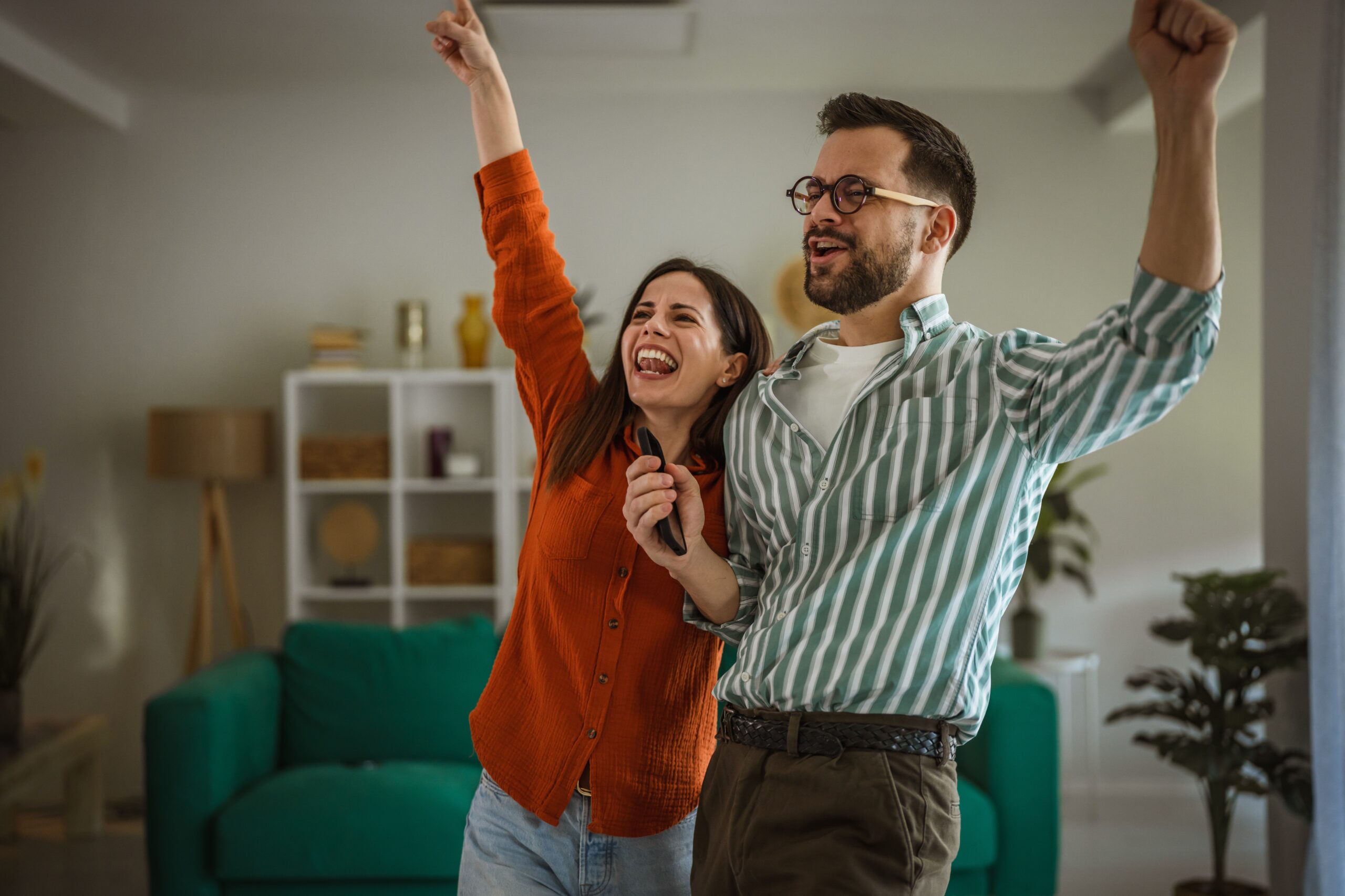 A woman and a man stand in a living room, smiling and cheering with their arms raised—celebrating peace of mind thanks to their Homeowners Insurance Bolingbrook. The room features a green sofa, decor-filled shelves, and a potted plant.