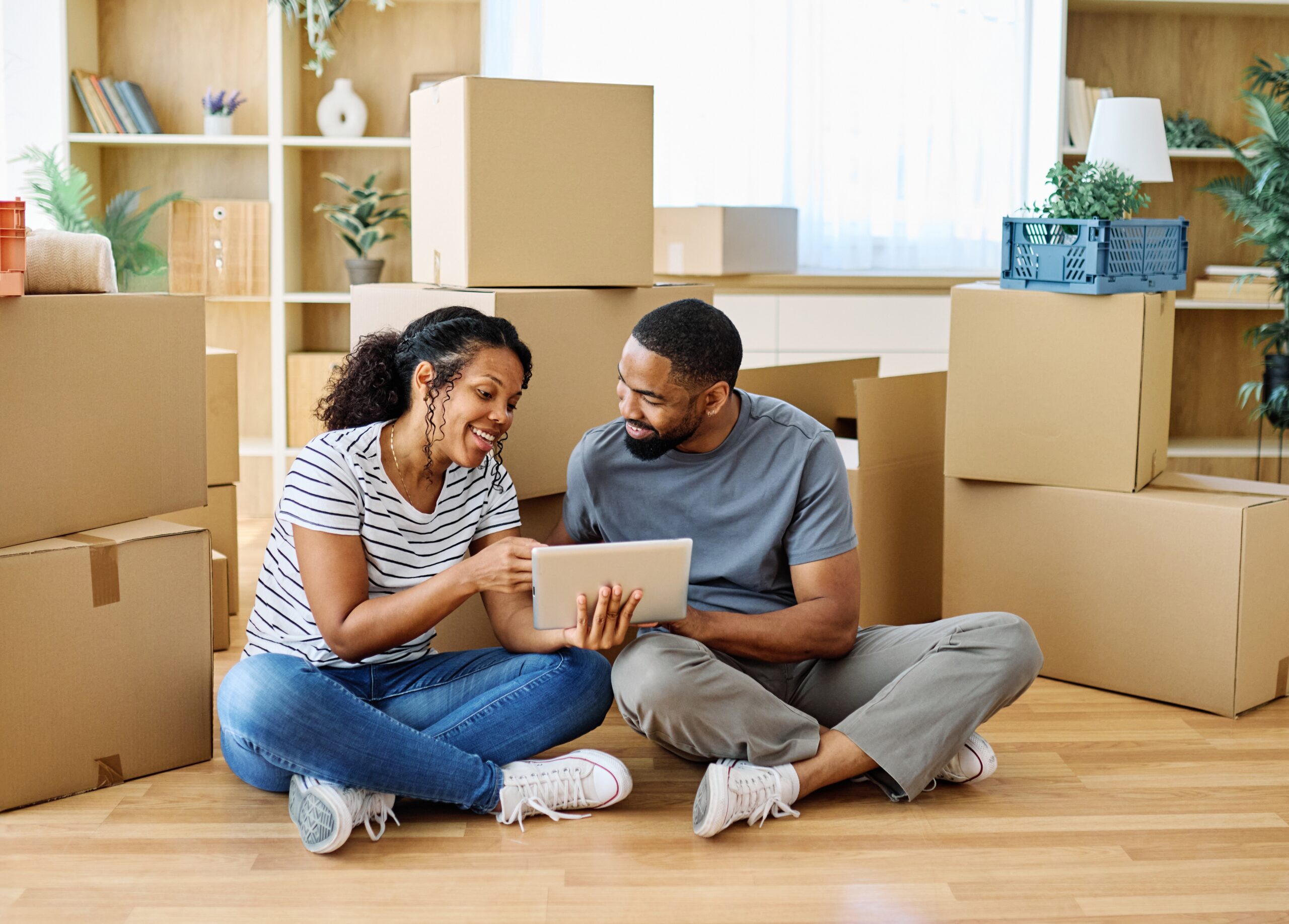 A man and woman sit on the floor surrounded by cardboard boxes, looking at a tablet together. With shelves, plants, and large windows in view, they appear to be moving in—perhaps researching Bolingbrook IL homeowners insurance for their new home.