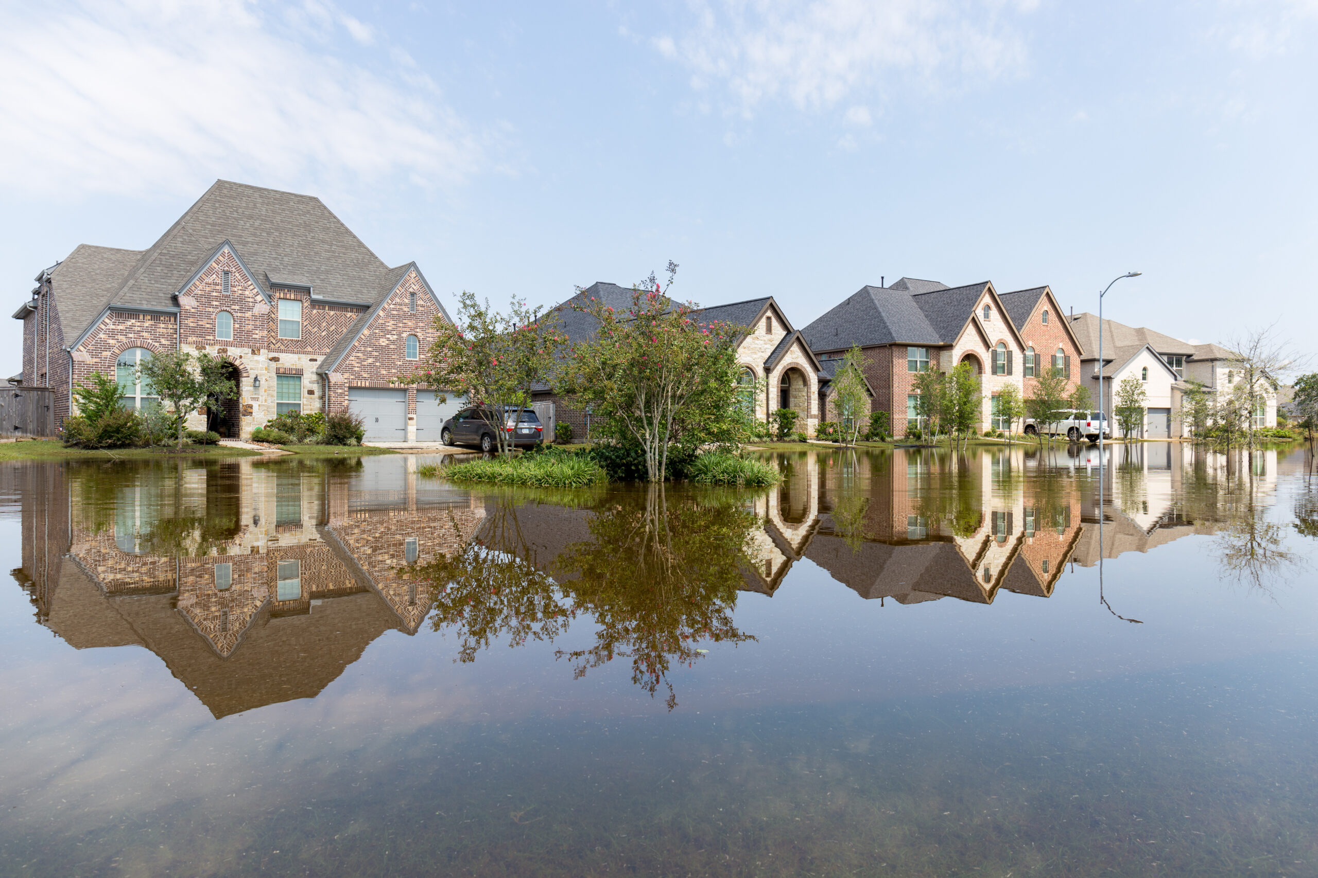 A row of suburban houses in Berwyn, partially surrounded by floodwater, with trees, parked vehicles, and reflections in the water under a partly cloudy sky—highlighting the importance of homeowners insurance Berwyn.