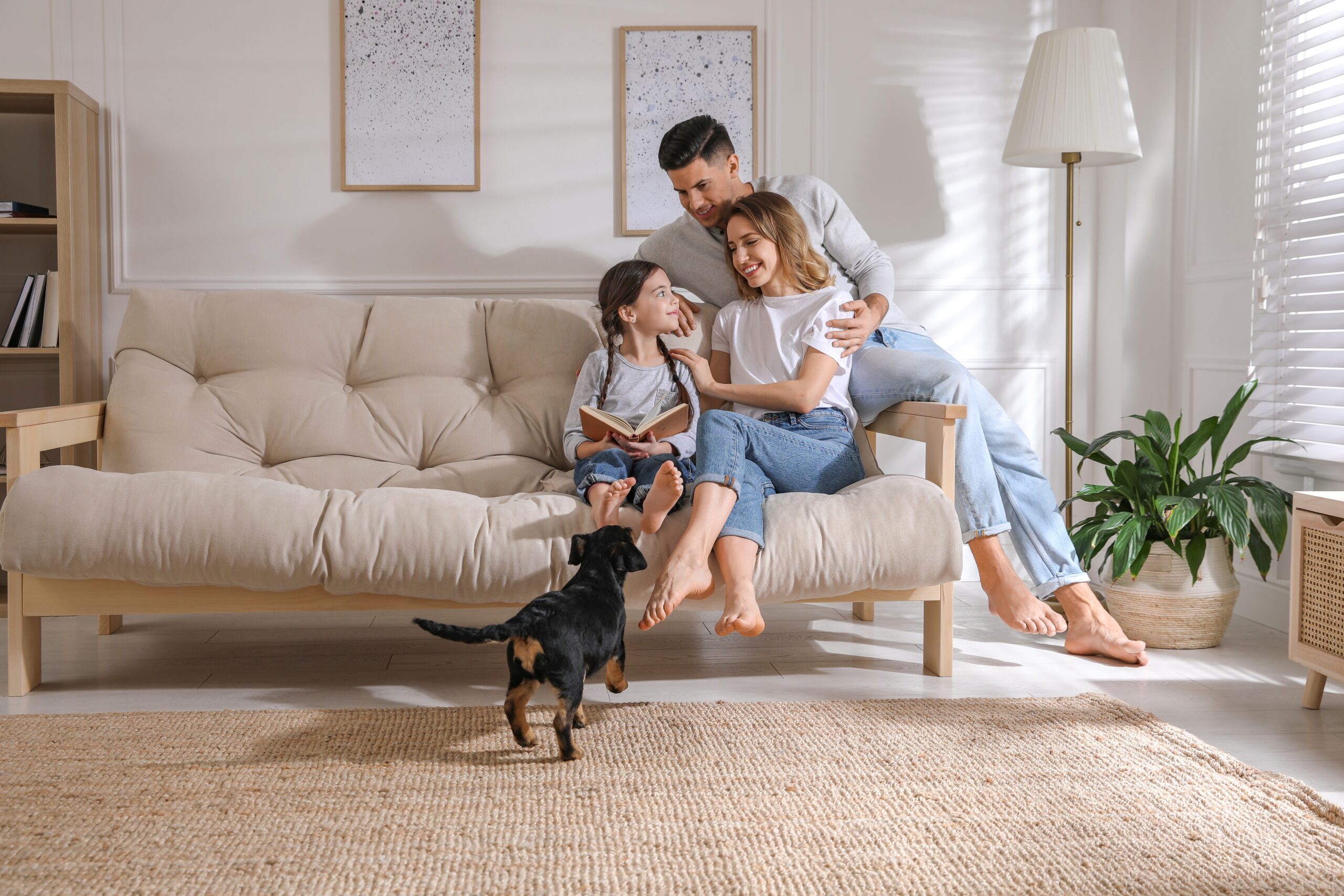 A man, woman, and young girl sit together on a beige sofa, smiling and talking while the girl holds a book. A small black dog stands on the rug in their bright, modern living room—a cozy setting for homeowners insurance Brookfield IL.