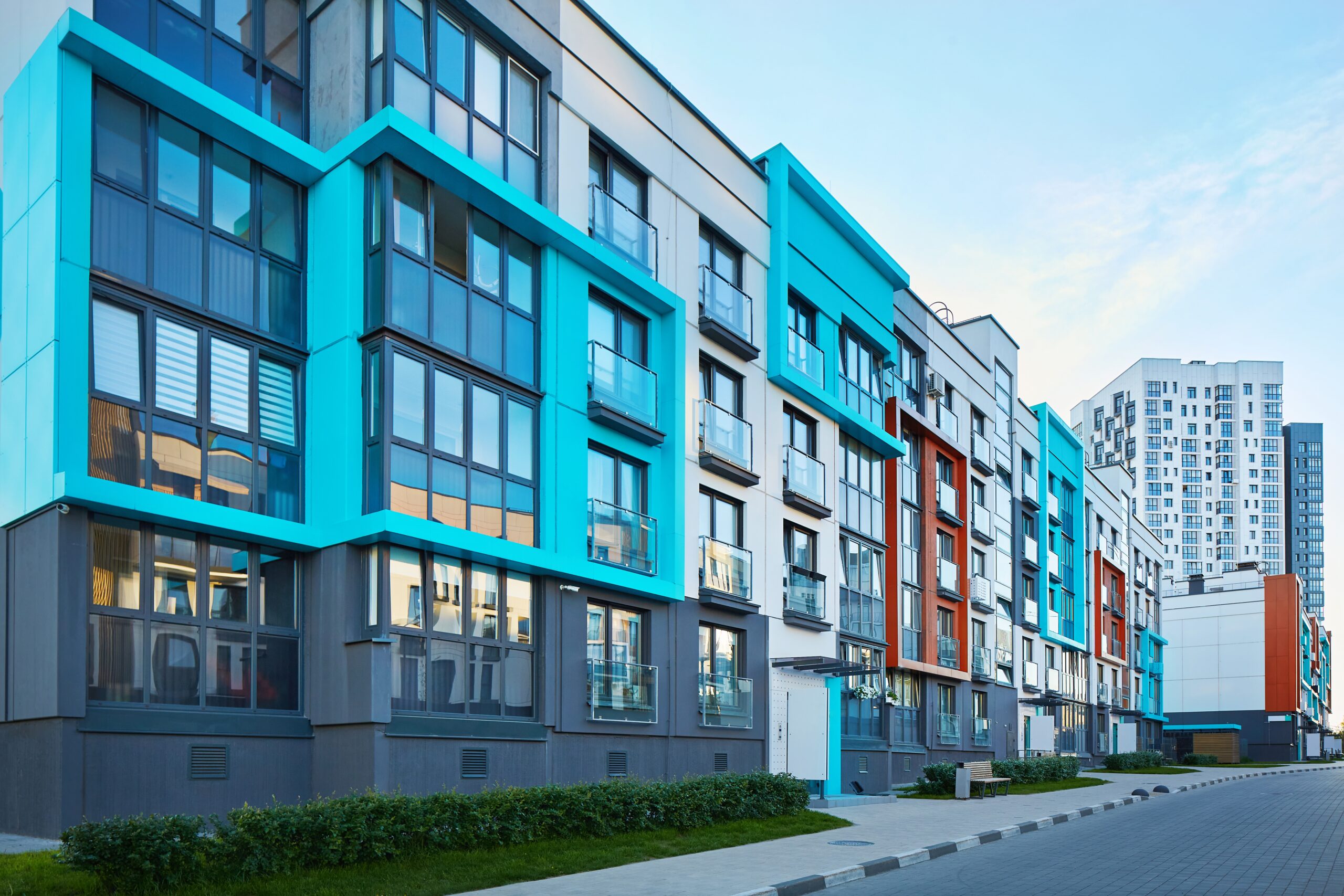 Modern apartment buildings with large windows and colorful geometric facades in blue, gray, and orange tones line a clean walkway with greenery—perfect for those seeking stylish living and peace of mind with homeowners insurance Arlington. Another high-rise stands in the background.