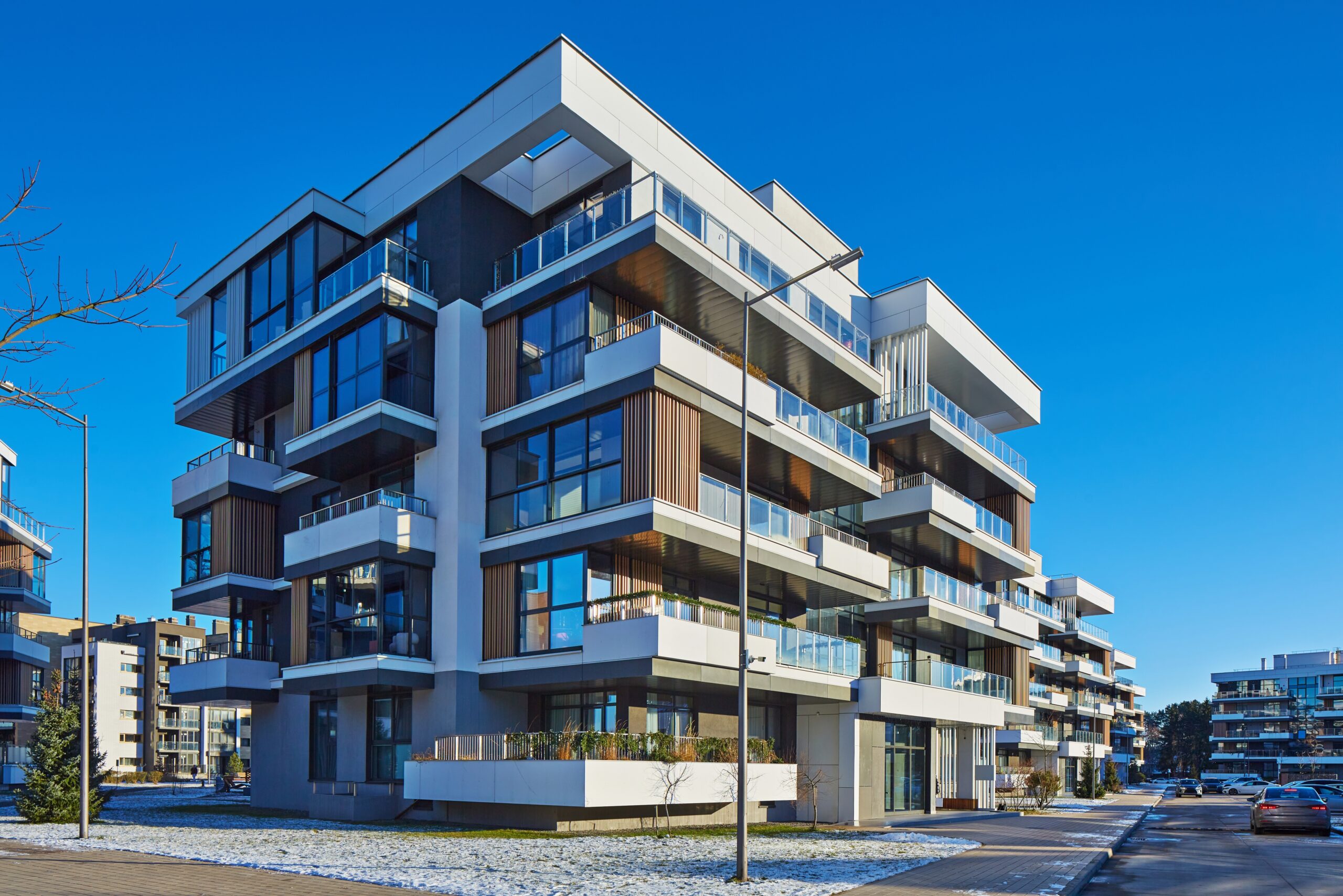 A modern, multi-story apartment building with large glass windows and balconies on each floor stands under a clear blue sky, snow on the ground, and a paved walkway in front—perfect for those seeking homeowners insurance Arlington residents trust.