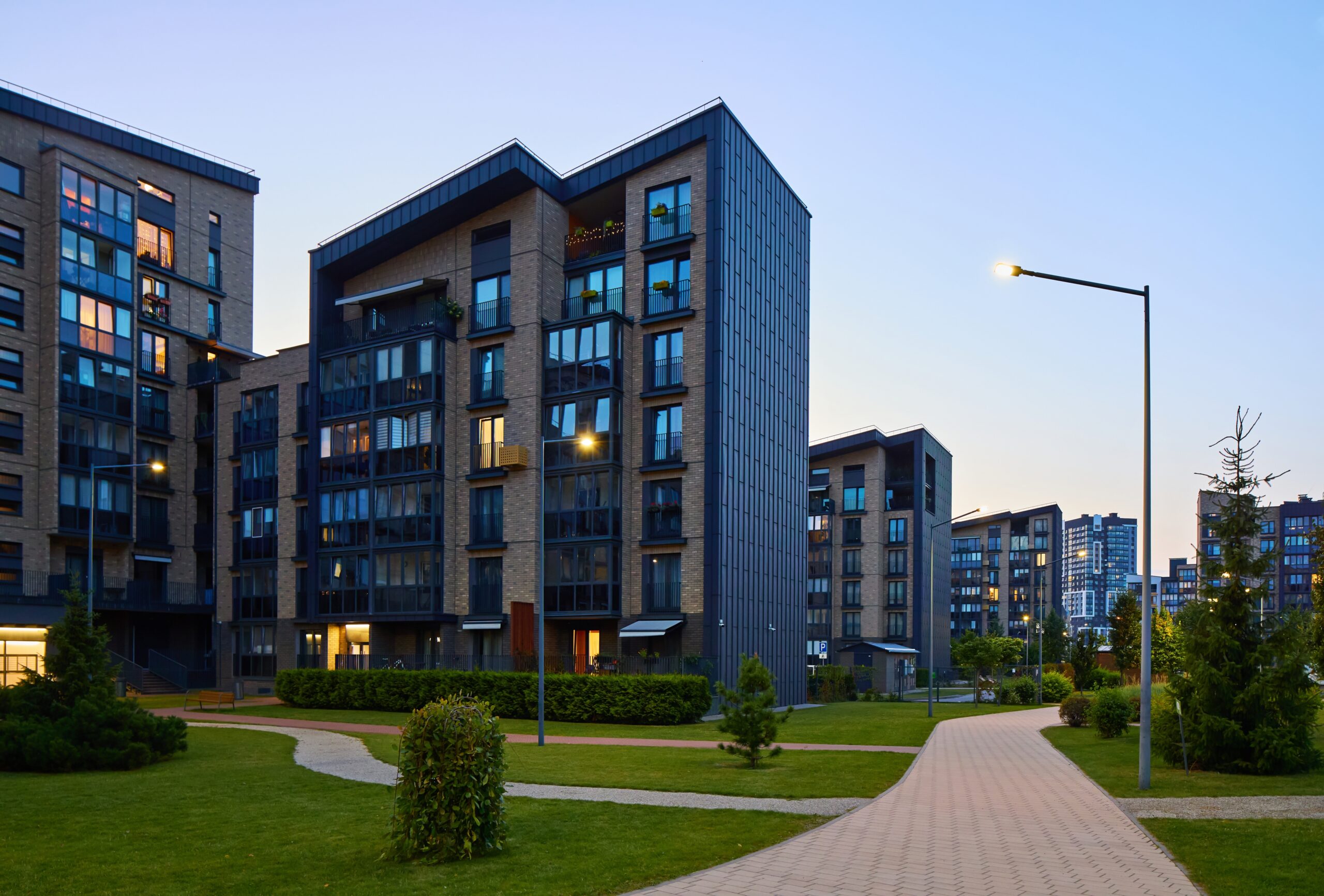Modern apartment buildings with multiple floors and large windows stand along a paved pathway, surrounded by green lawns and bushes under a clear early evening sky with streetlights turned on—perfect for residents seeking Addison homeowners insurance.