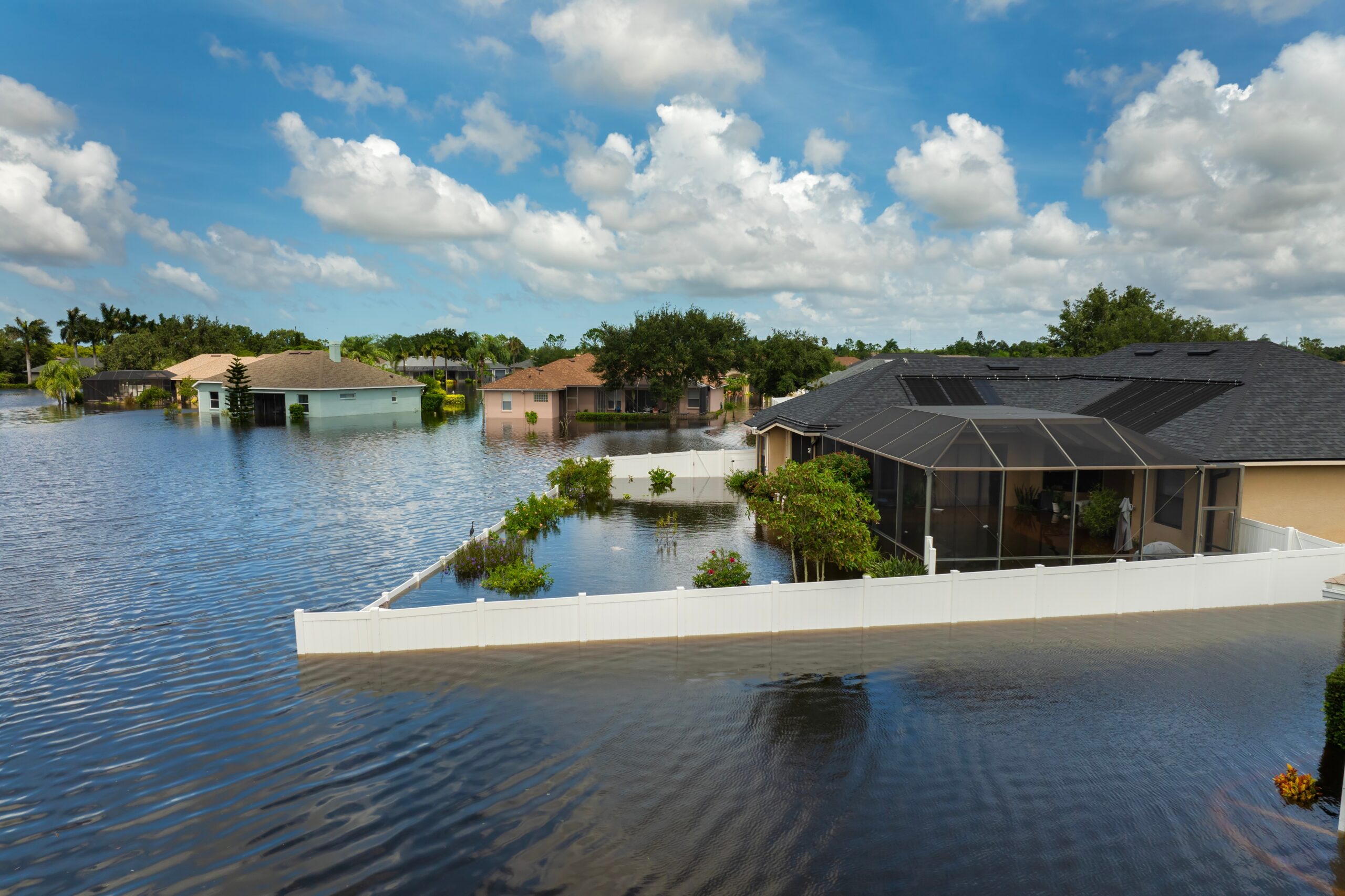 Residential neighborhood flooded with water, partially submerging homes, yards, and fencing under a blue sky with scattered clouds. Trees and shrubs rise above the floodwater—reminding residents of the importance of homeowners insurance Arlington.