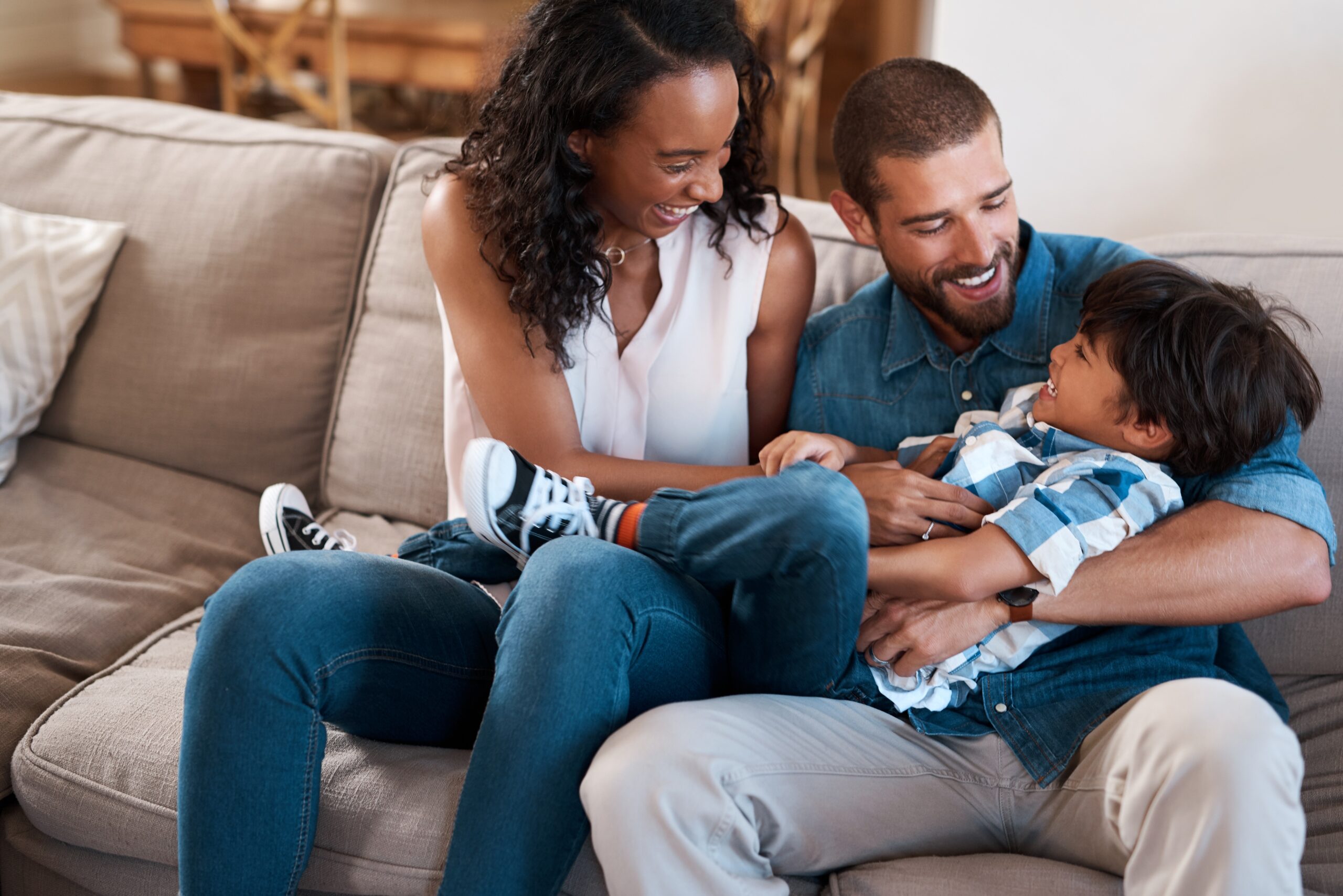 A woman and a man sit on a couch, smiling as they play with a young boy sprawled across their laps. The boy is laughing, and all three appear happy and relaxed—enjoying peace of mind thanks to reliable homeowners insurance Arlington families trust.