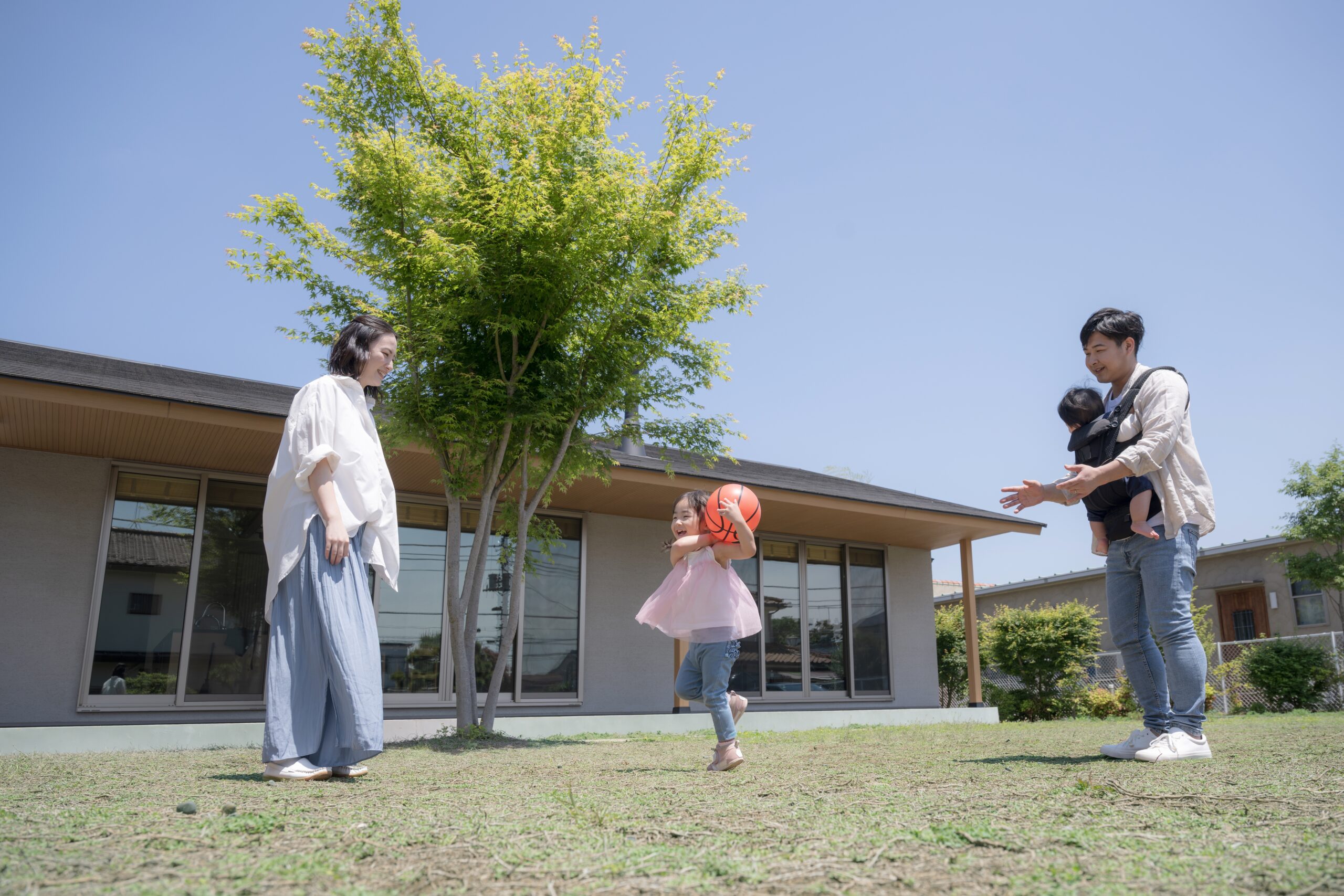 A woman, a man holding a child, and a young girl with a red ball are outside on a grassy area near their house on a sunny day—an ideal scene for families protected by Addison homeowners insurance. The adults watch as the girl walks toward them.