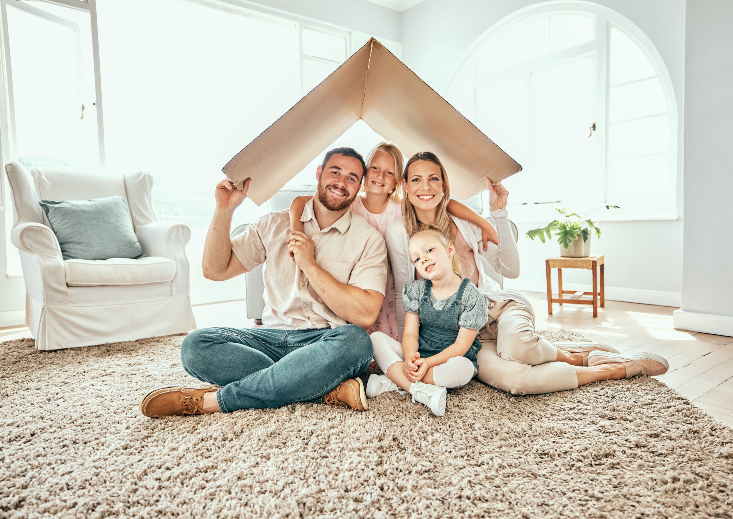 A man, woman, and two young girls sit on a carpeted floor in a bright room, holding a cardboard roof over their heads and smiling—capturing the comfort homeowners insurance Arlington can help protect. A white armchair and small table with a plant are in the background.