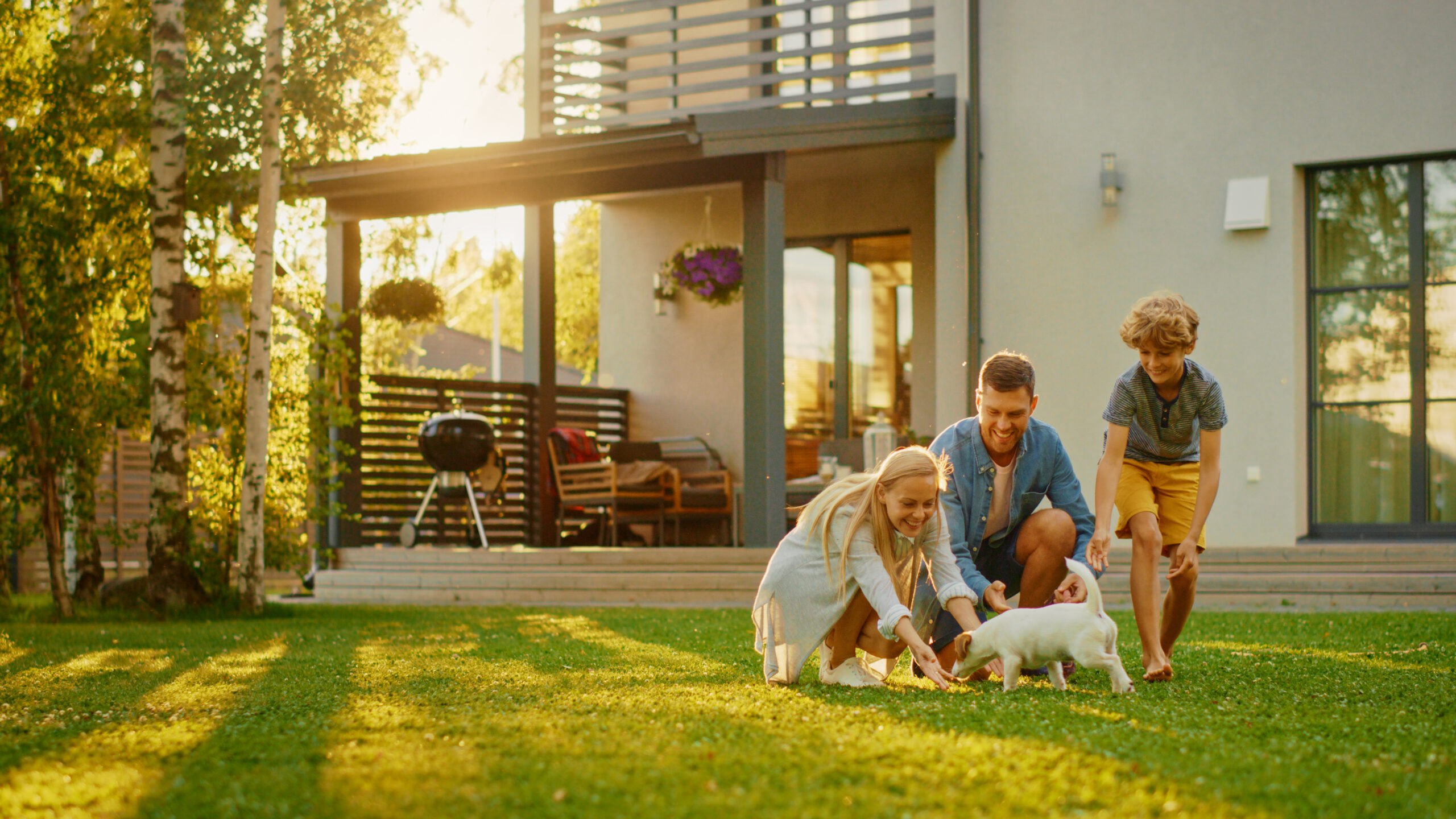 A family of three and their small dog play together on a grassy lawn in front of a modern house, while outdoor furniture and a barbecue grill sit on the porch—an idyllic scene for those with Addison homeowners insurance peace of mind.