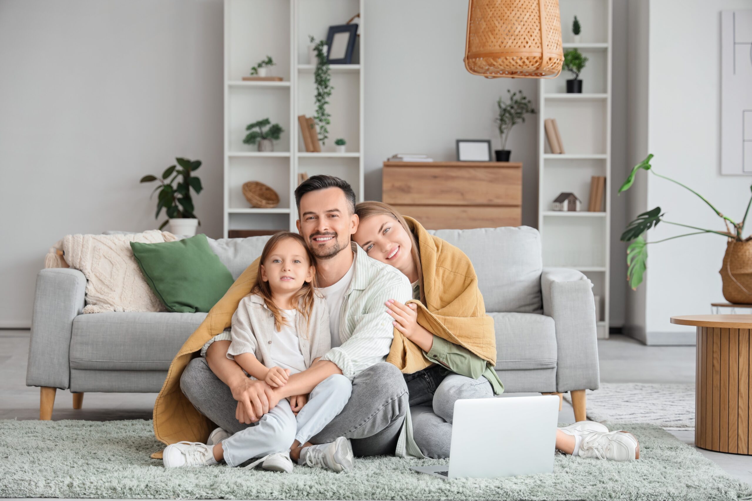 A man, woman, and child sit closely together on a living room floor, wrapped in a blanket. Smiling at a laptop, this happy family enjoys peace of mind with homeowners insurance in Brookfield, IL. A gray sofa, plants, and shelves fill the background.