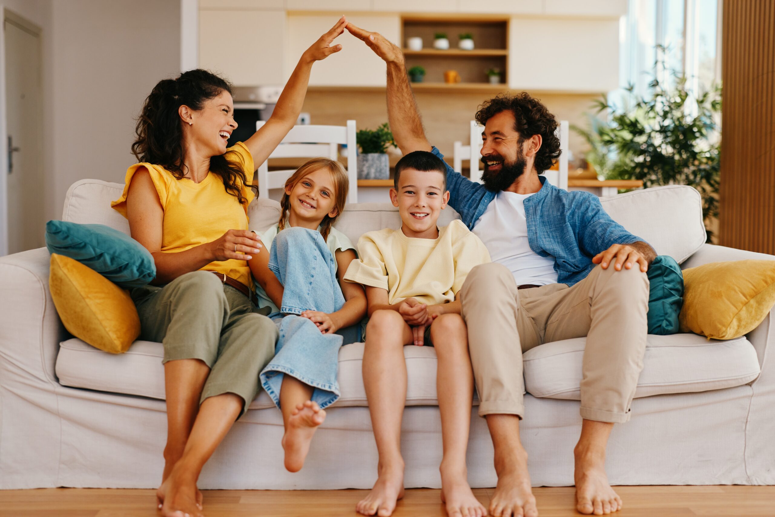 A woman and man sit on a couch with two children between them, smiling. The adults raise their hands to form an arch above the children, capturing the warmth and security that comes with homeowners insurance Arlington families trust.