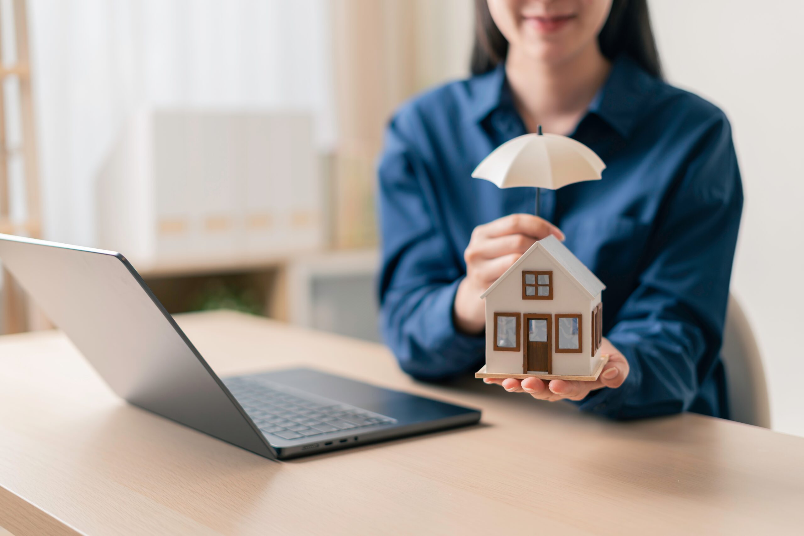 A woman in a blue shirt sits at a desk with a laptop, holding a model house and shielding it with an umbrella—representing the protection and peace of mind offered by Addison homeowners insurance.