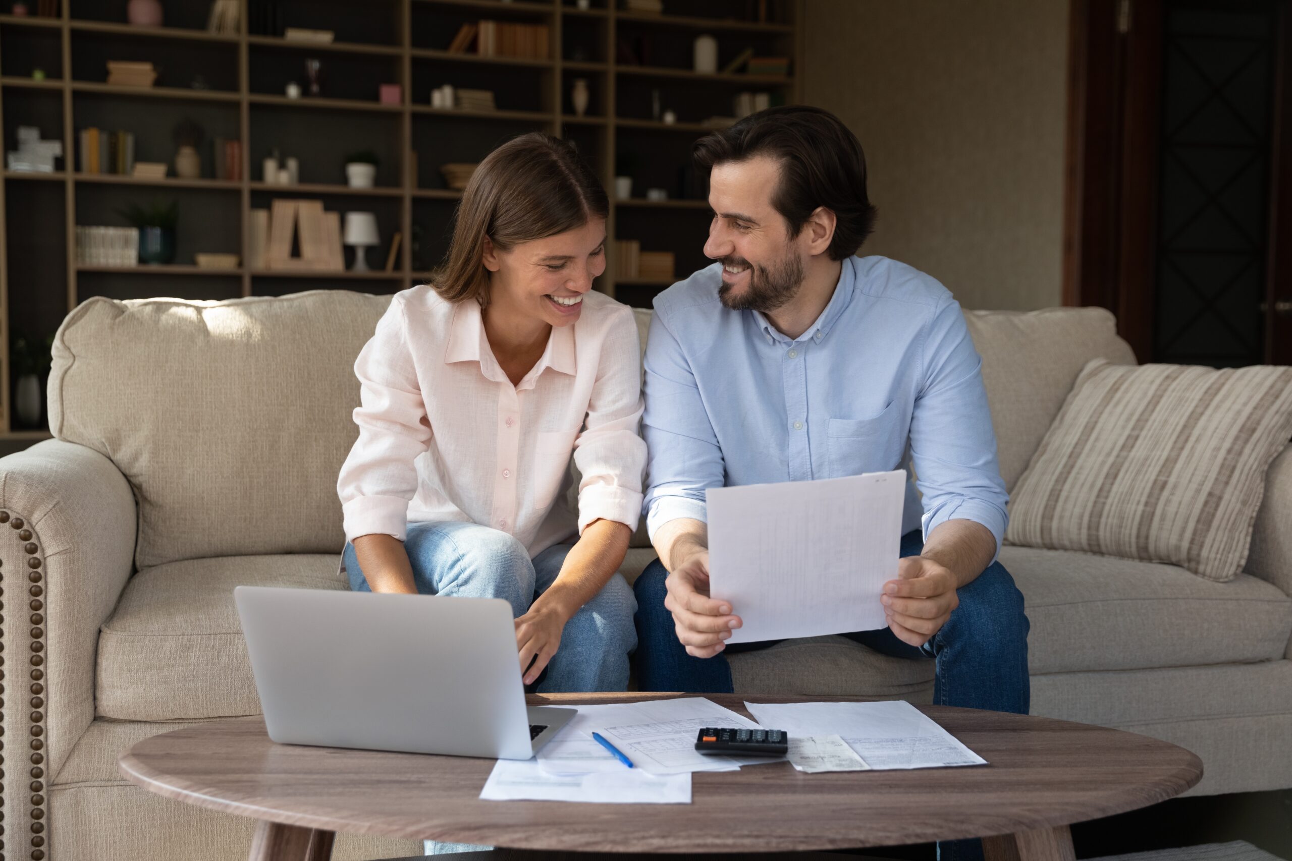 A man and woman sit on a couch looking at paperwork, with a laptop, calculator, and documents on the coffee table in front of them. They appear to be reviewing financial information, possibly related to Addison homeowners insurance.