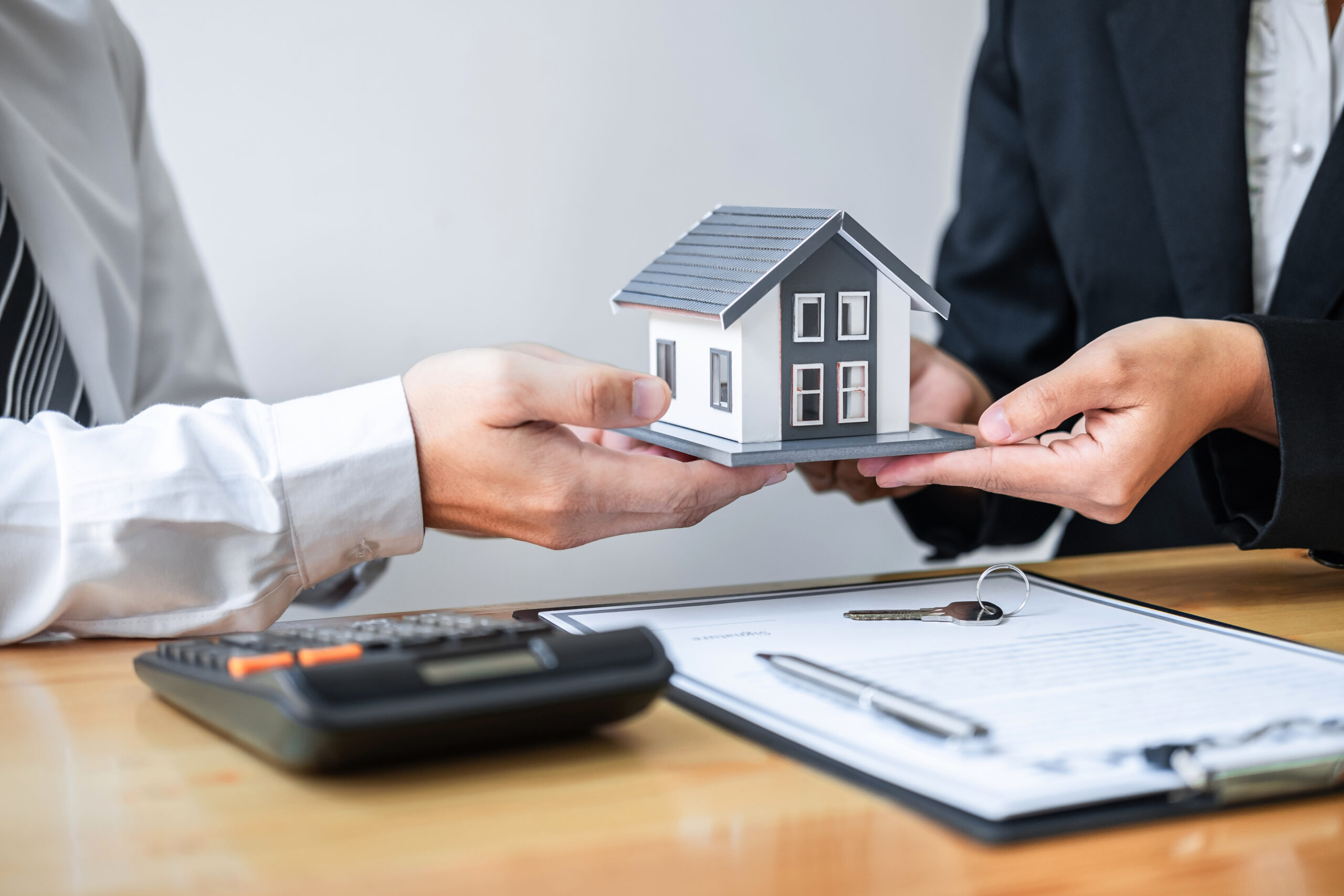 Two people in business attire exchange a model house over a desk with a contract, pen, calculator, and an addison homeowners insurance document, suggesting a real estate transaction or agreement.