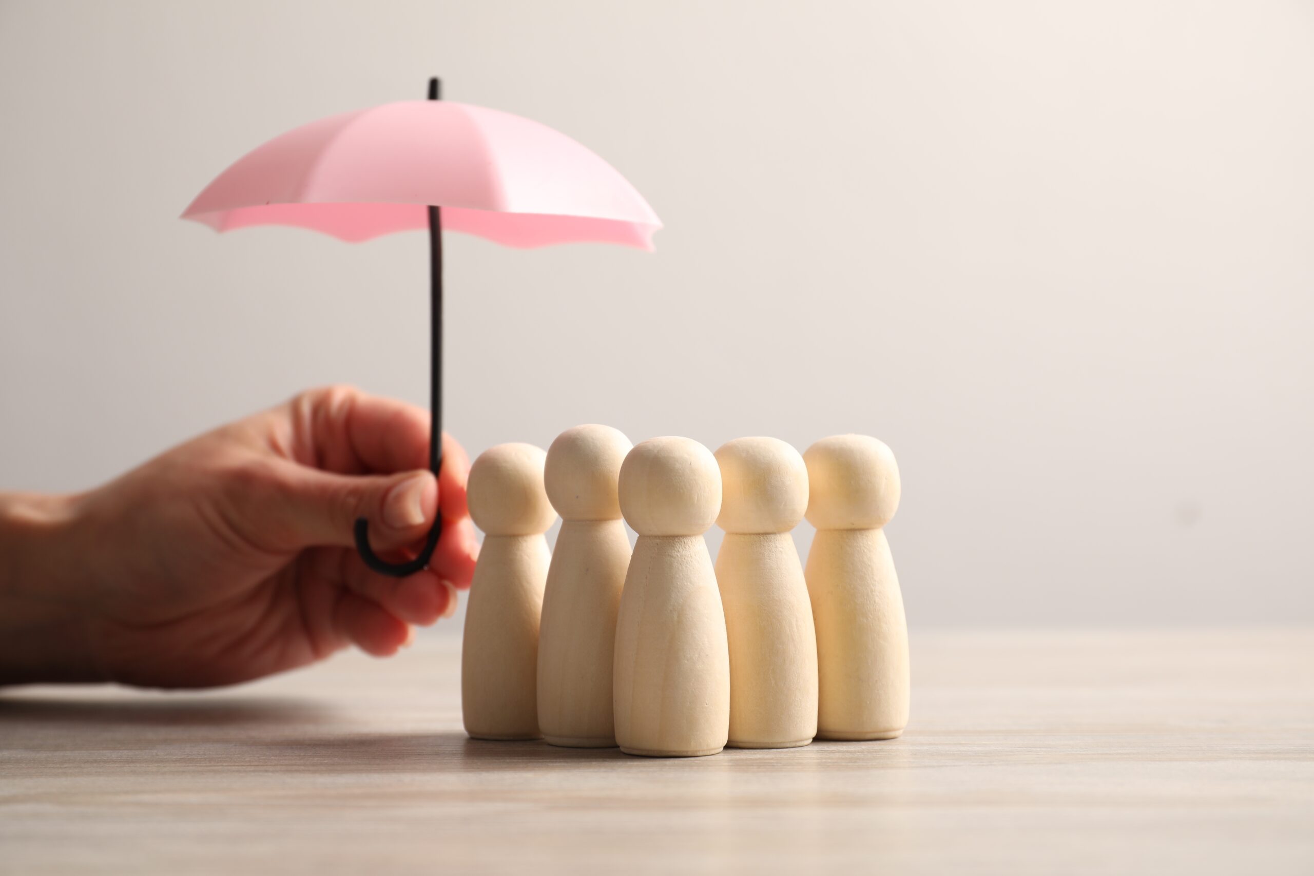 A hand holds a small pink umbrella over six wooden figurines standing in a row on a light-colored surface, symbolizing the protection offered by homeowners insurance in Brookfield, IL.