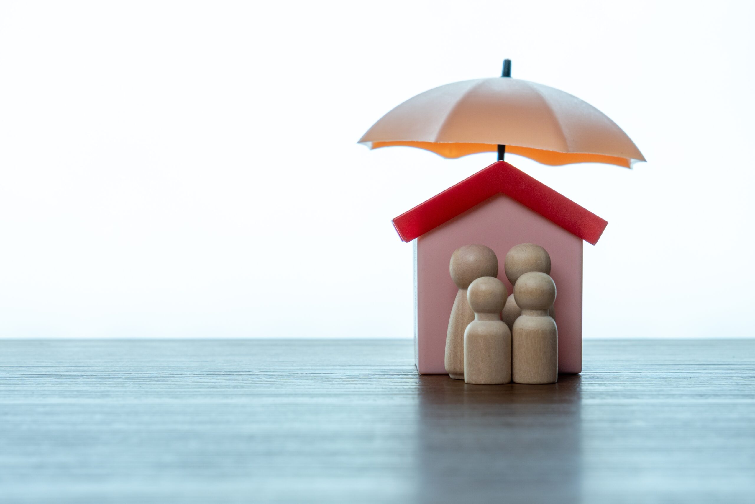 A small model house with four wooden human figurines in front, all under an orange umbrella, sits on a wooden surface—illustrating the protection addison homeowners insurance provides—against a plain white background.