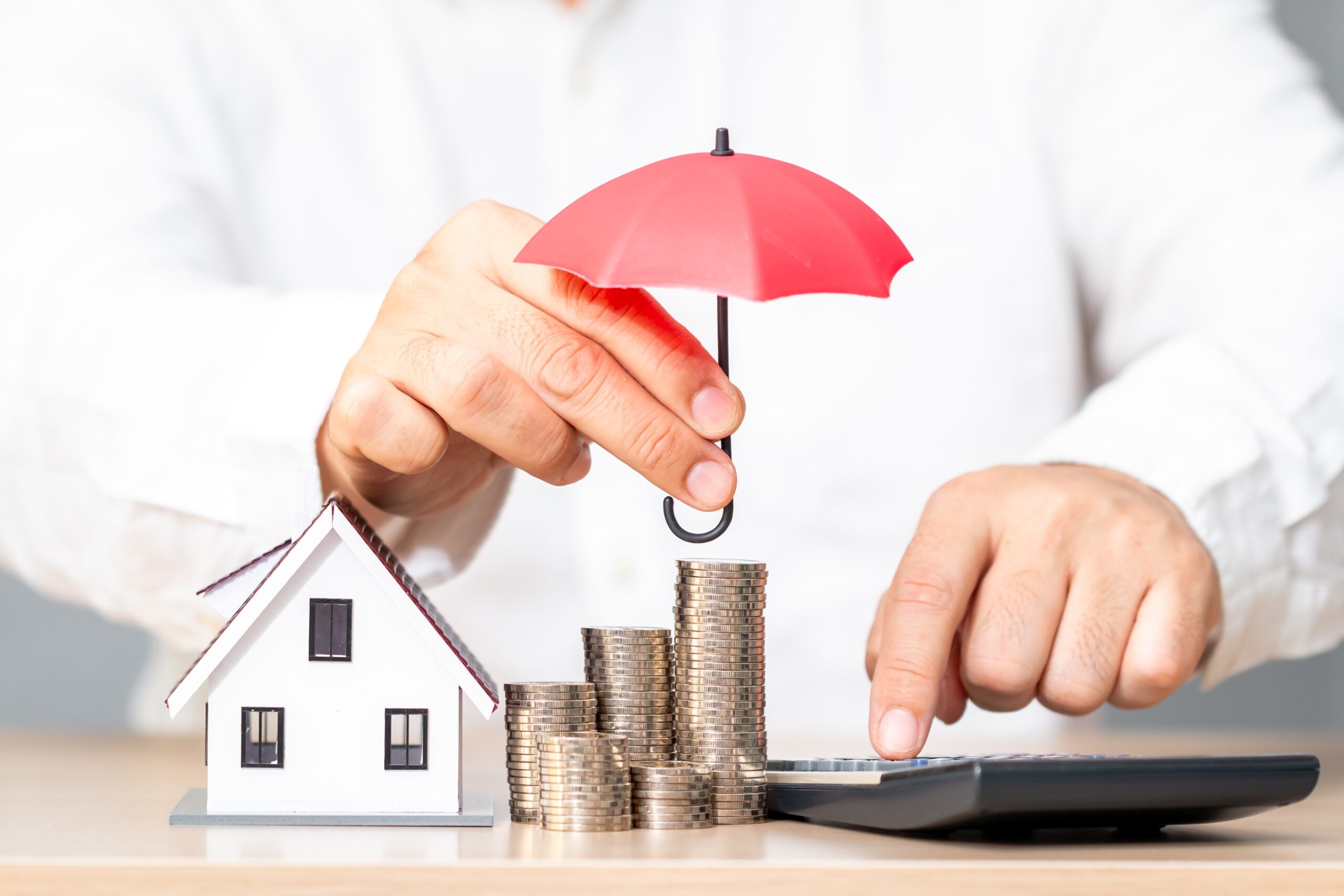 A person holds a small red umbrella over stacks of coins next to a model house, pressing calculator buttons—symbolizing financial protection or homeowners insurance Arlington residents can trust.